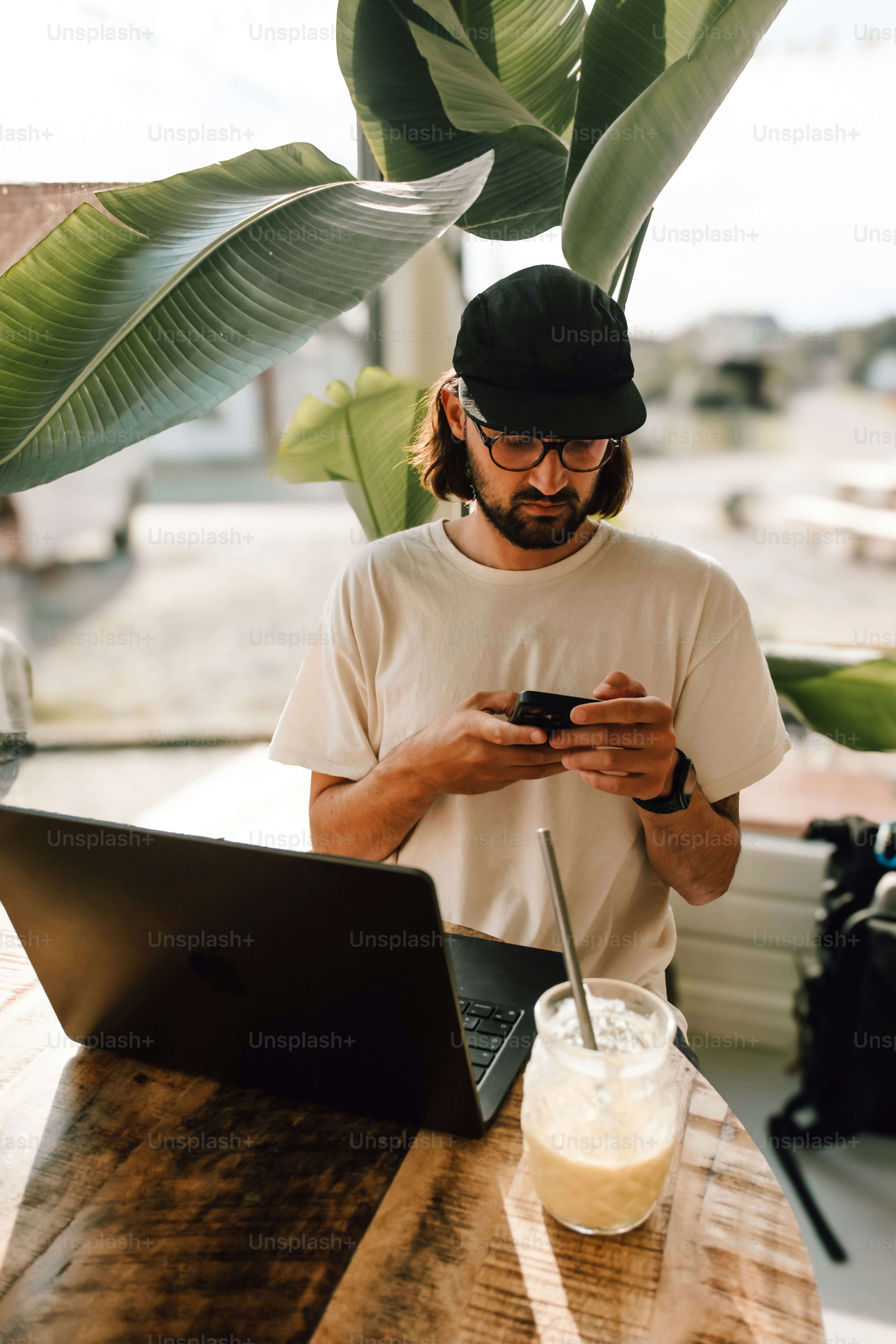 Man in cap using phone with laptop and drink.
