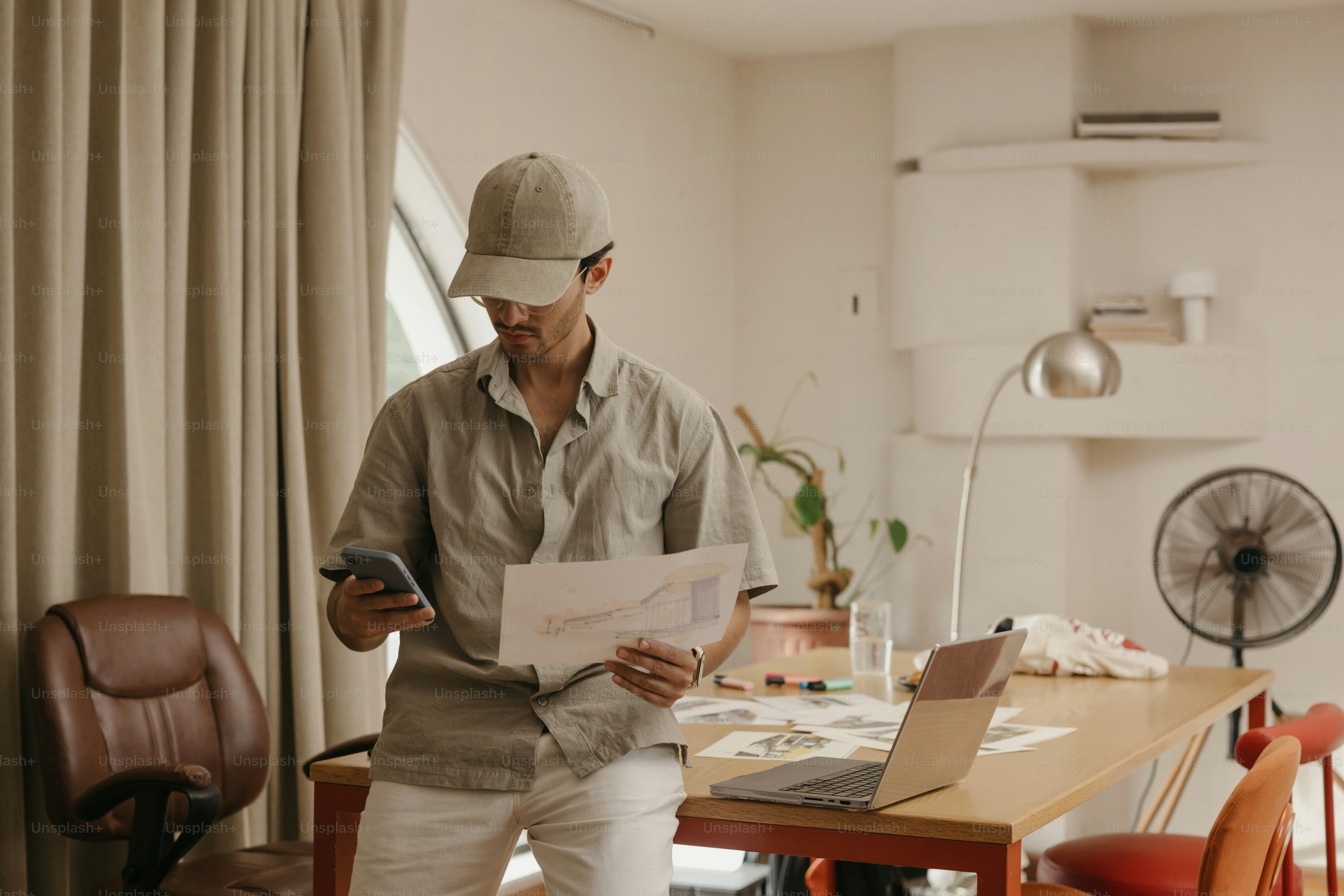 Man in cap checks phone and papers at desk.