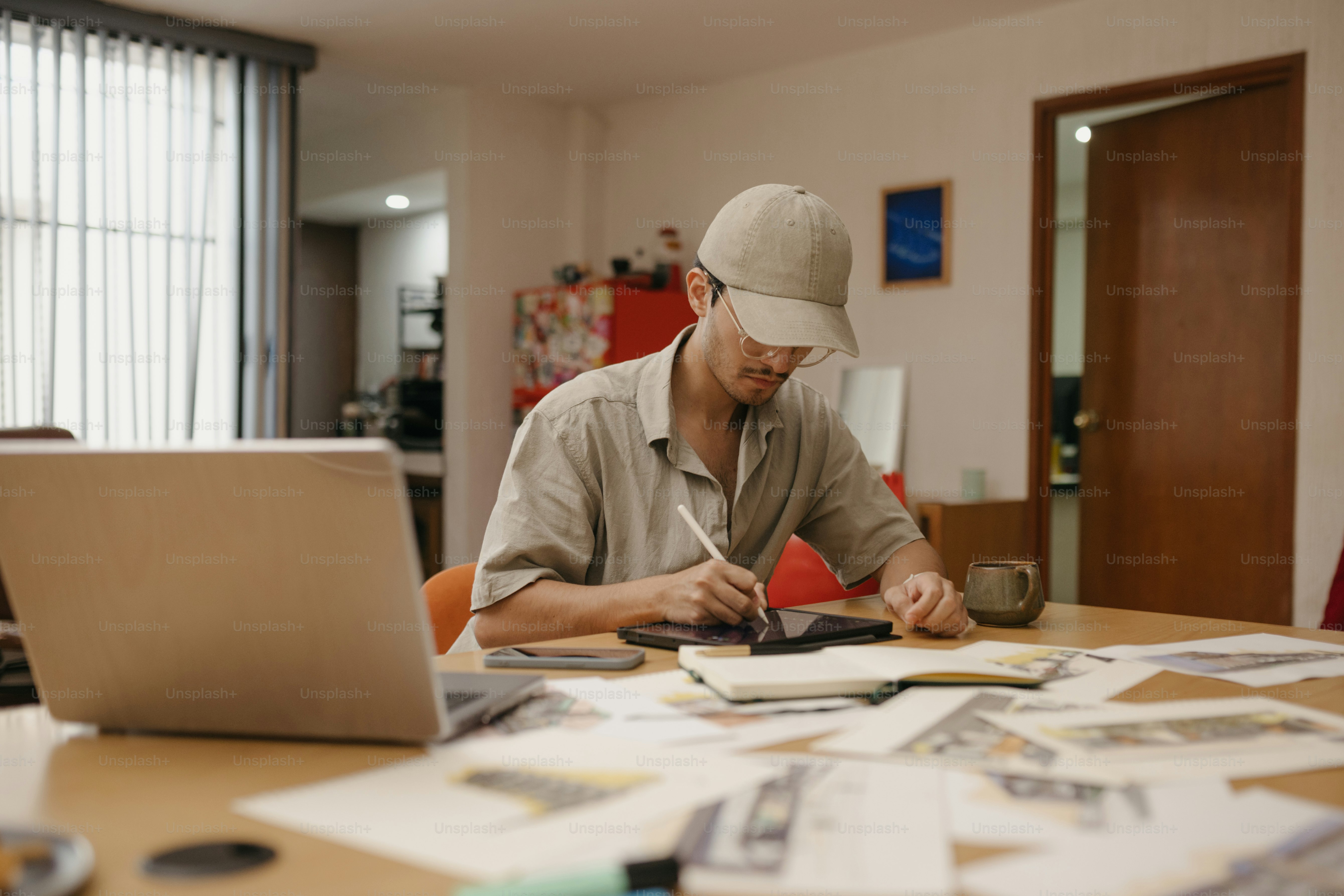 Man in baseball cap working at desk with laptop.