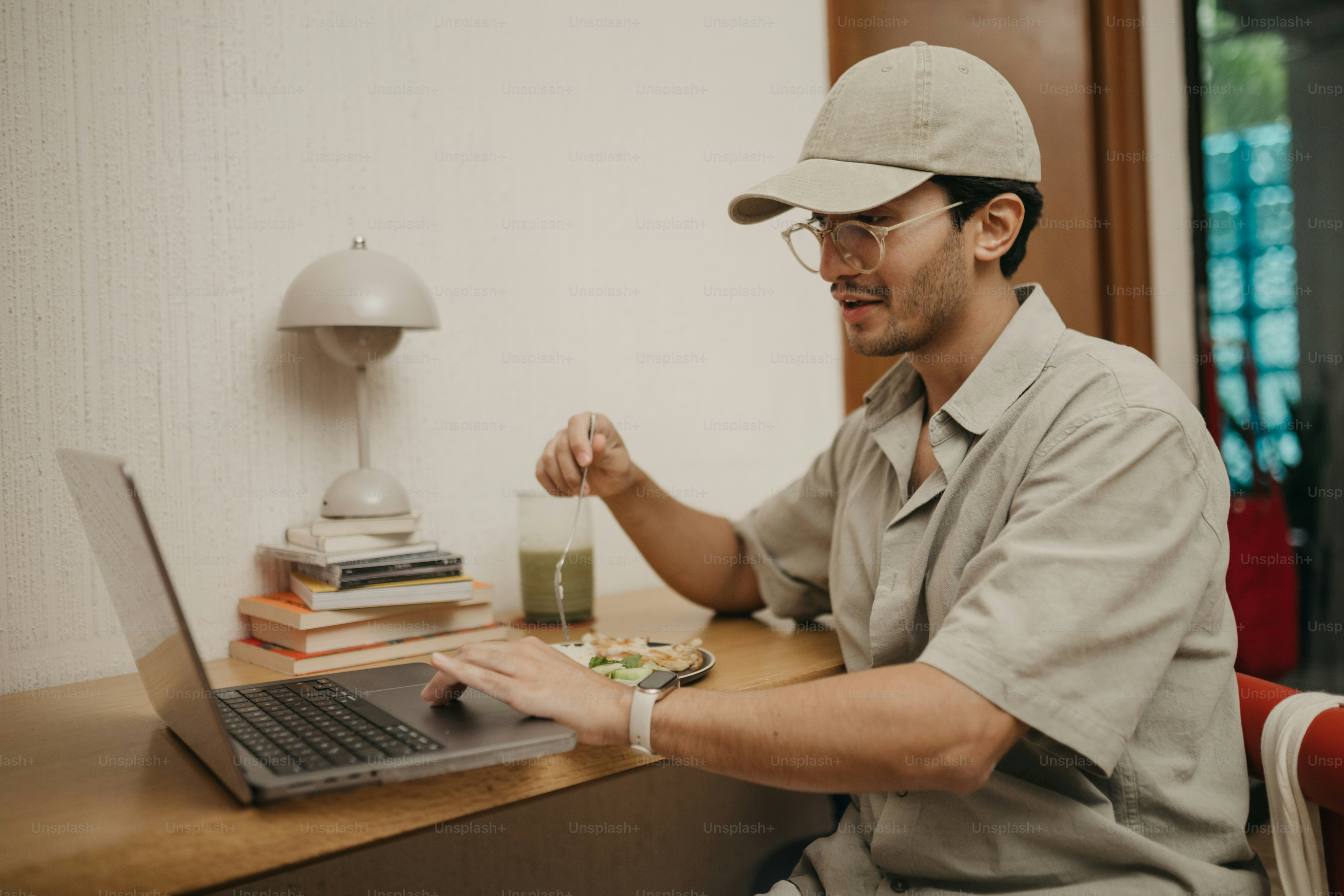 Man in cap and glasses working on laptop at desk.
