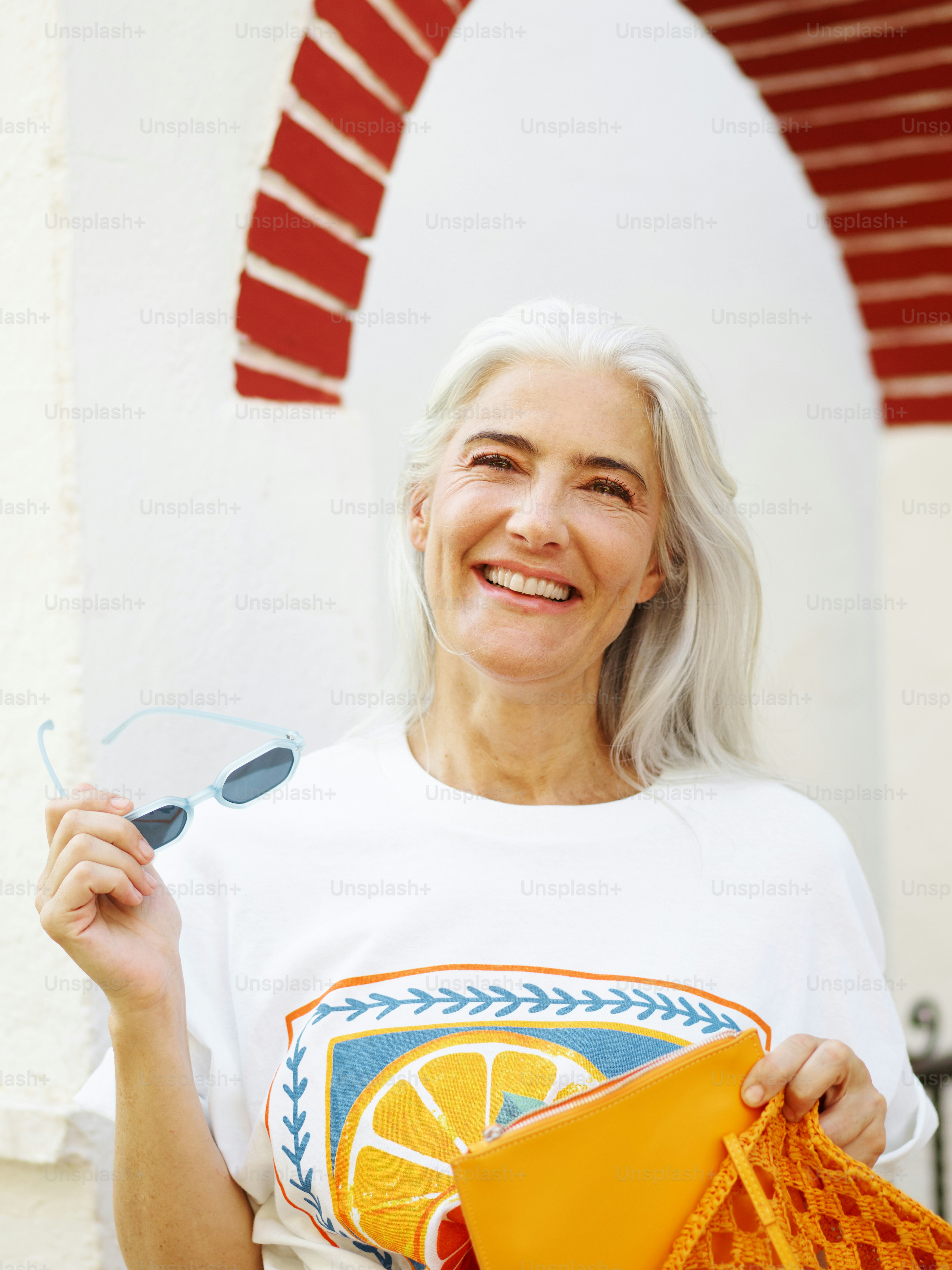 Smiling woman with gray hair holds sunglasses and bag.
