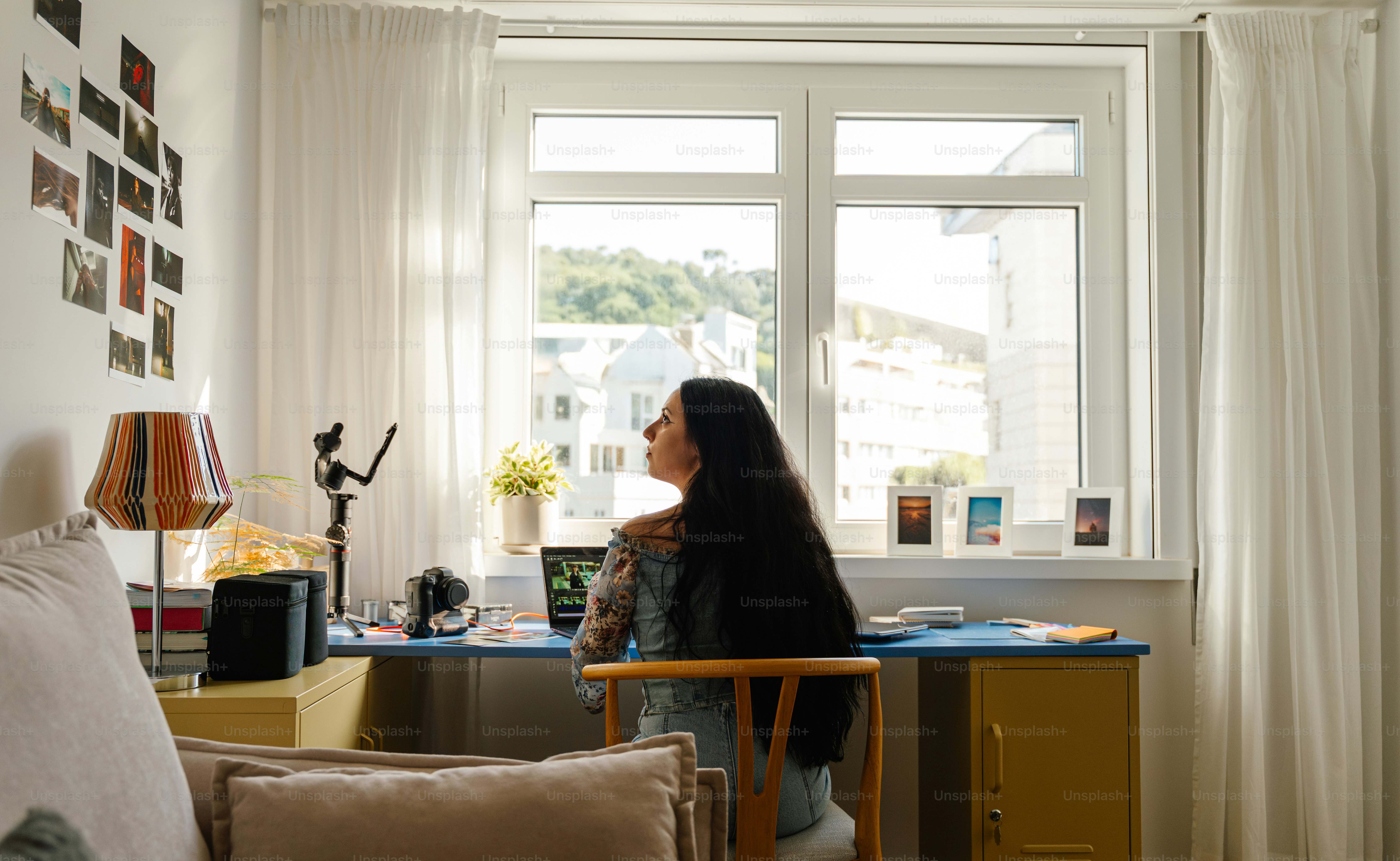 Woman sitting at desk by window with photos on wall.