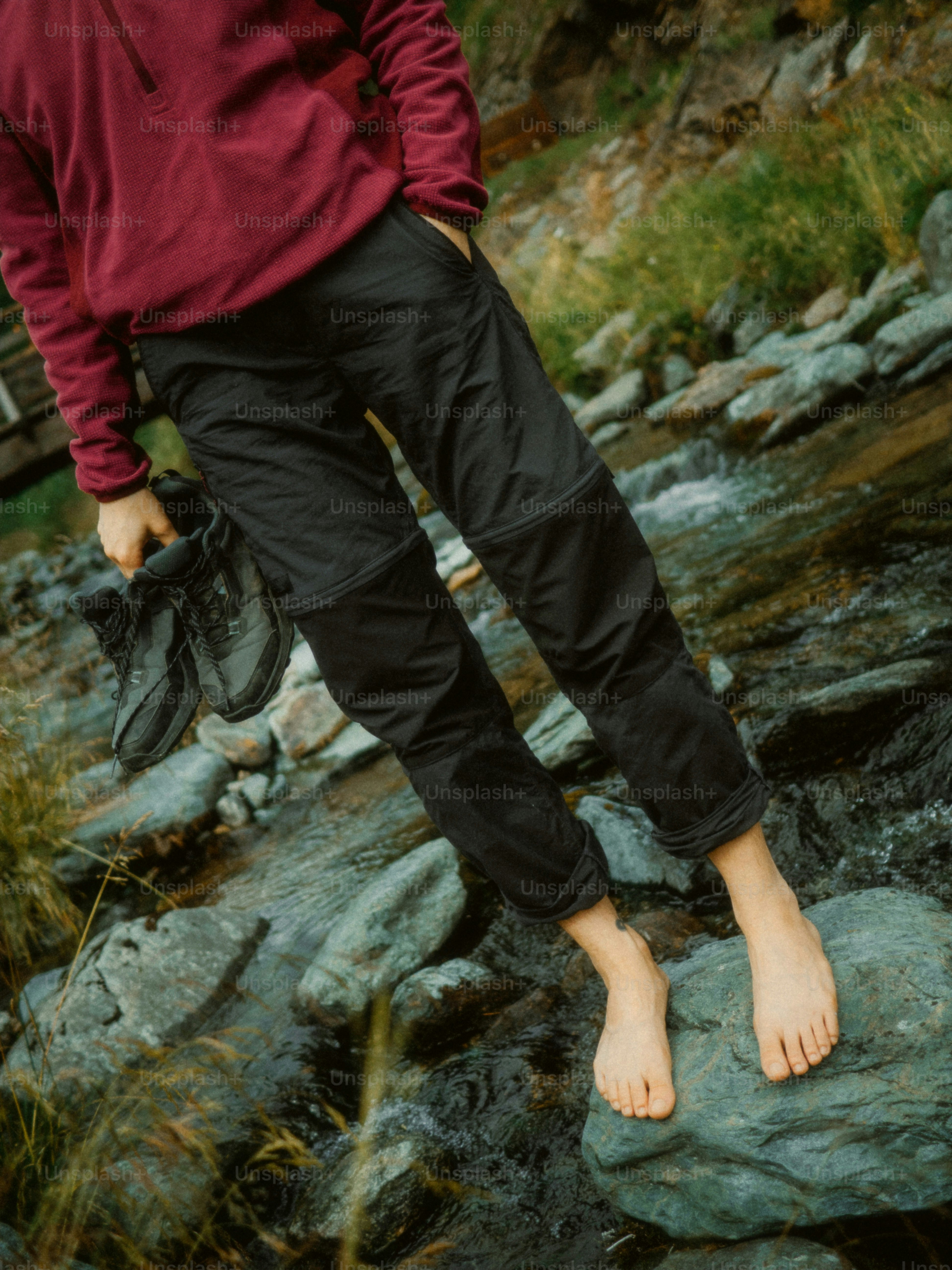 Barefoot person holding boots by a rocky stream
