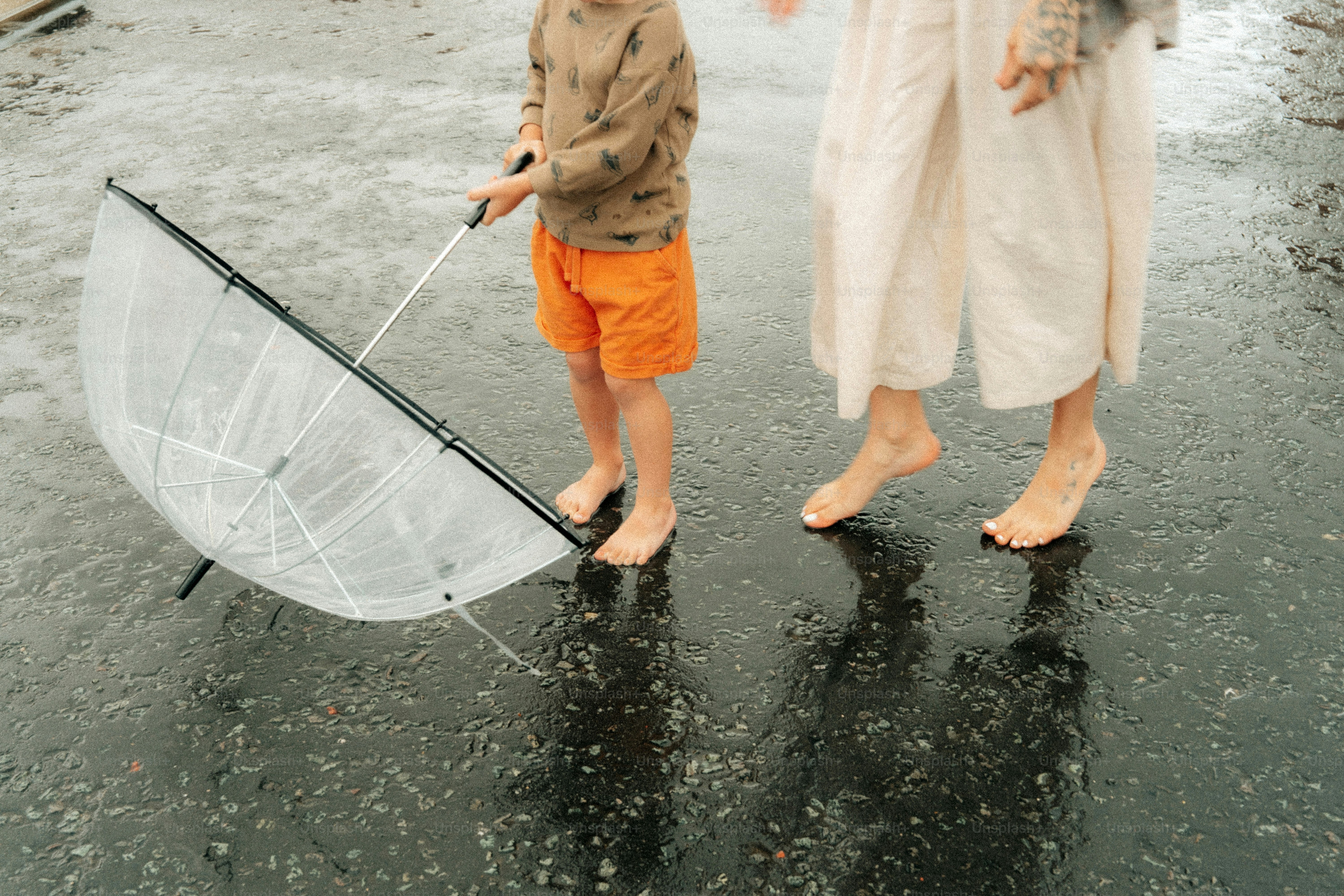 Barefoot child and adult holding umbrella in rain.