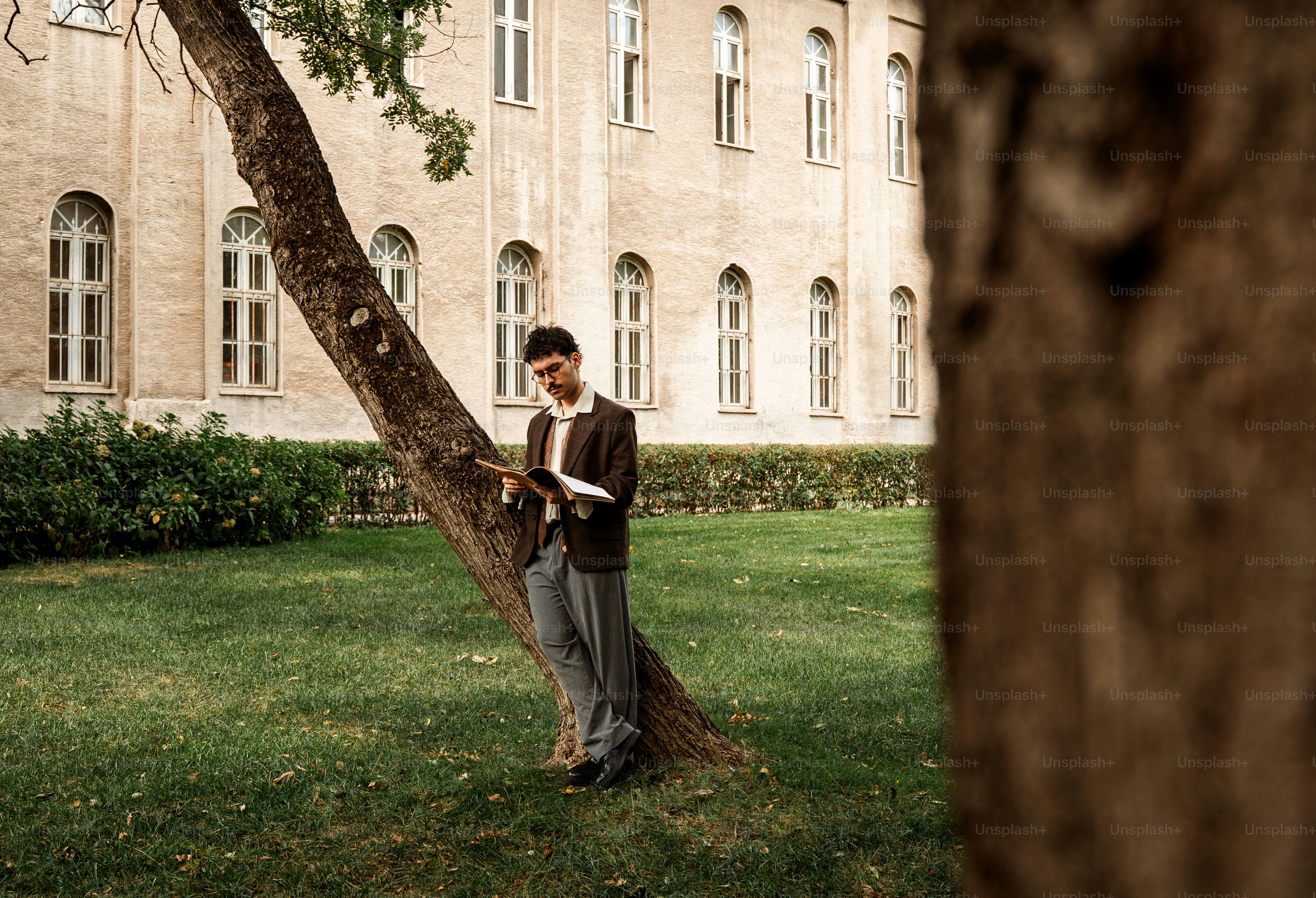 Young man reading a book leaning against a tree.