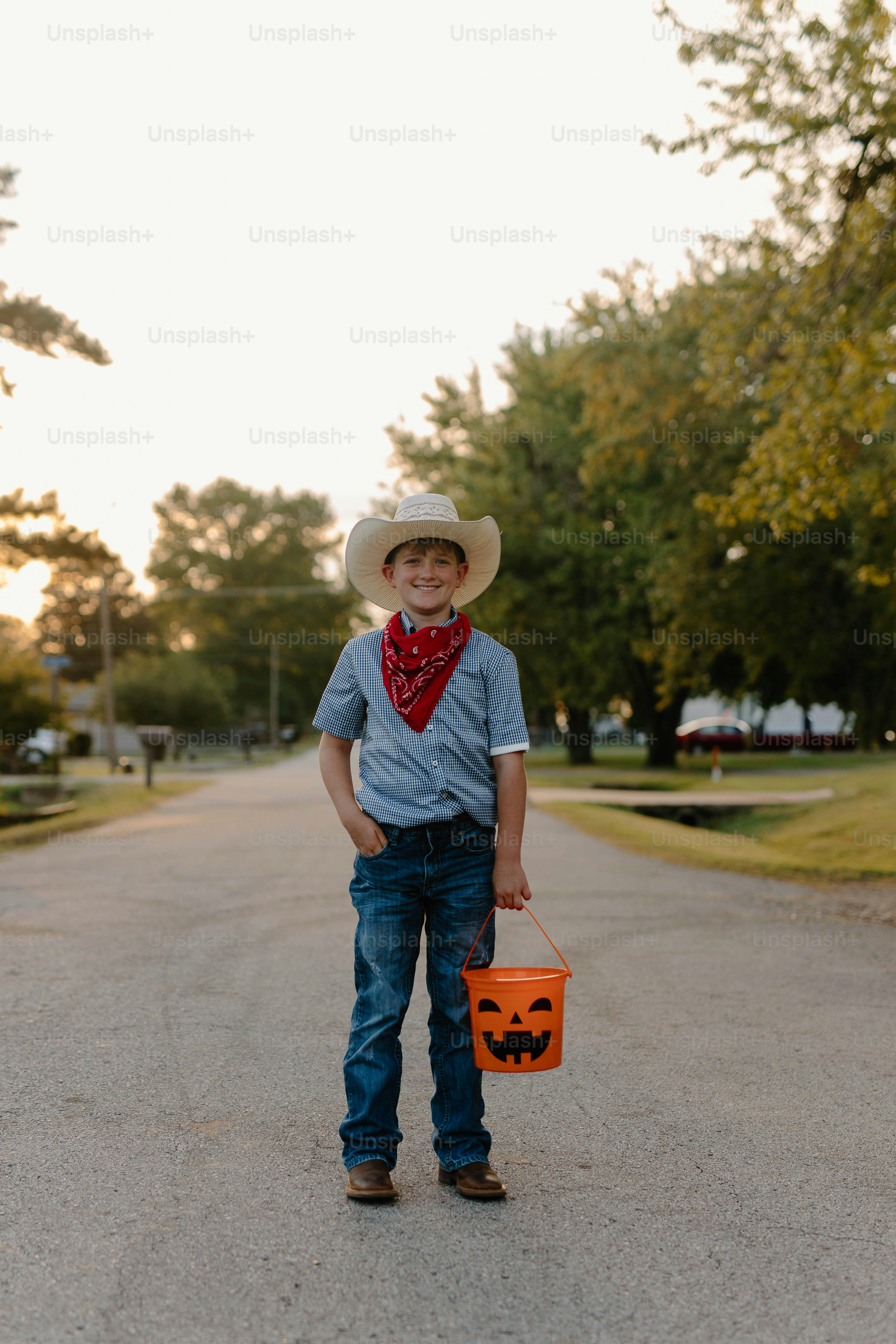 Boy in cowboy hat holds pumpkin bucket on street
