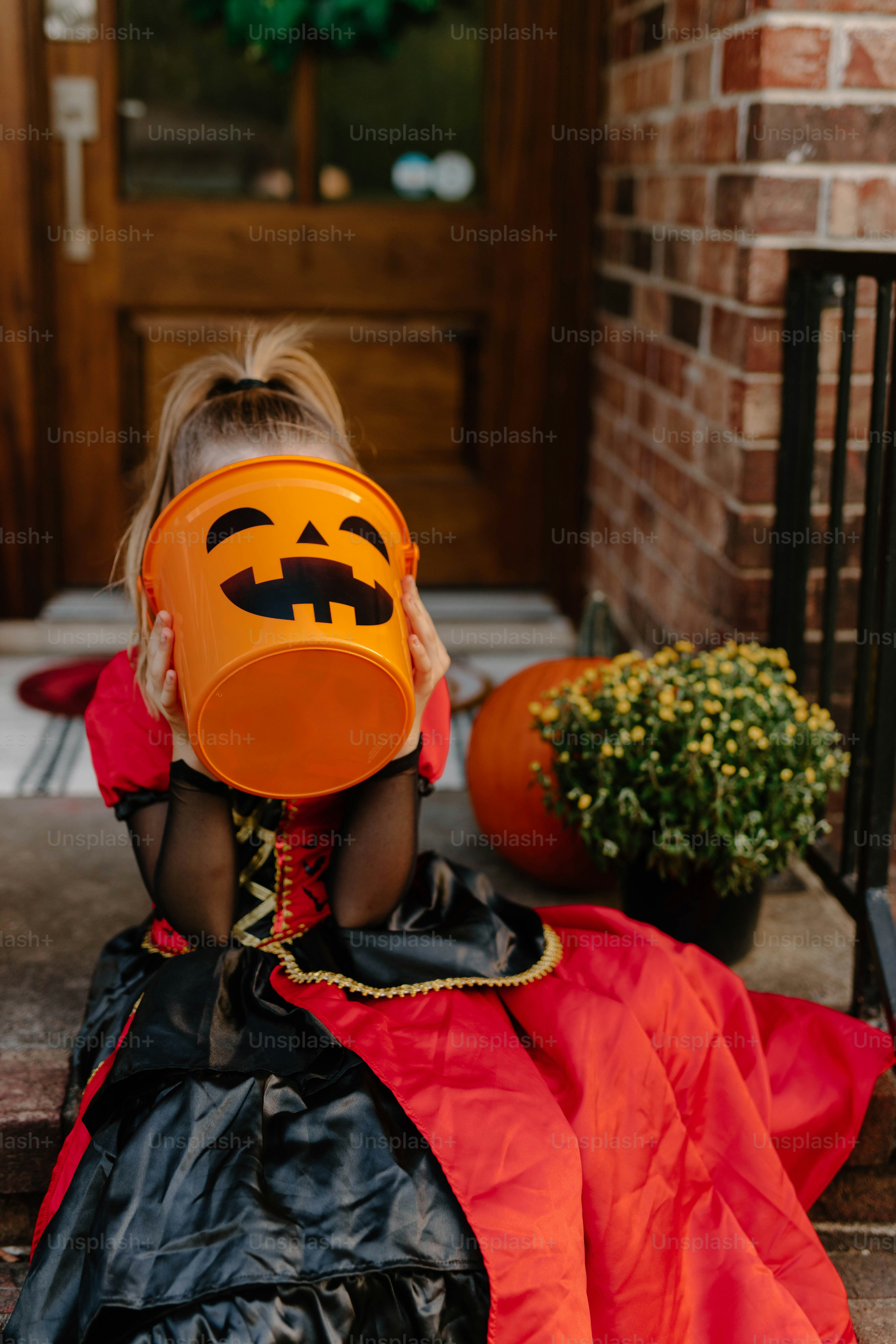 Child in costume holds pumpkin bucket on porch.
