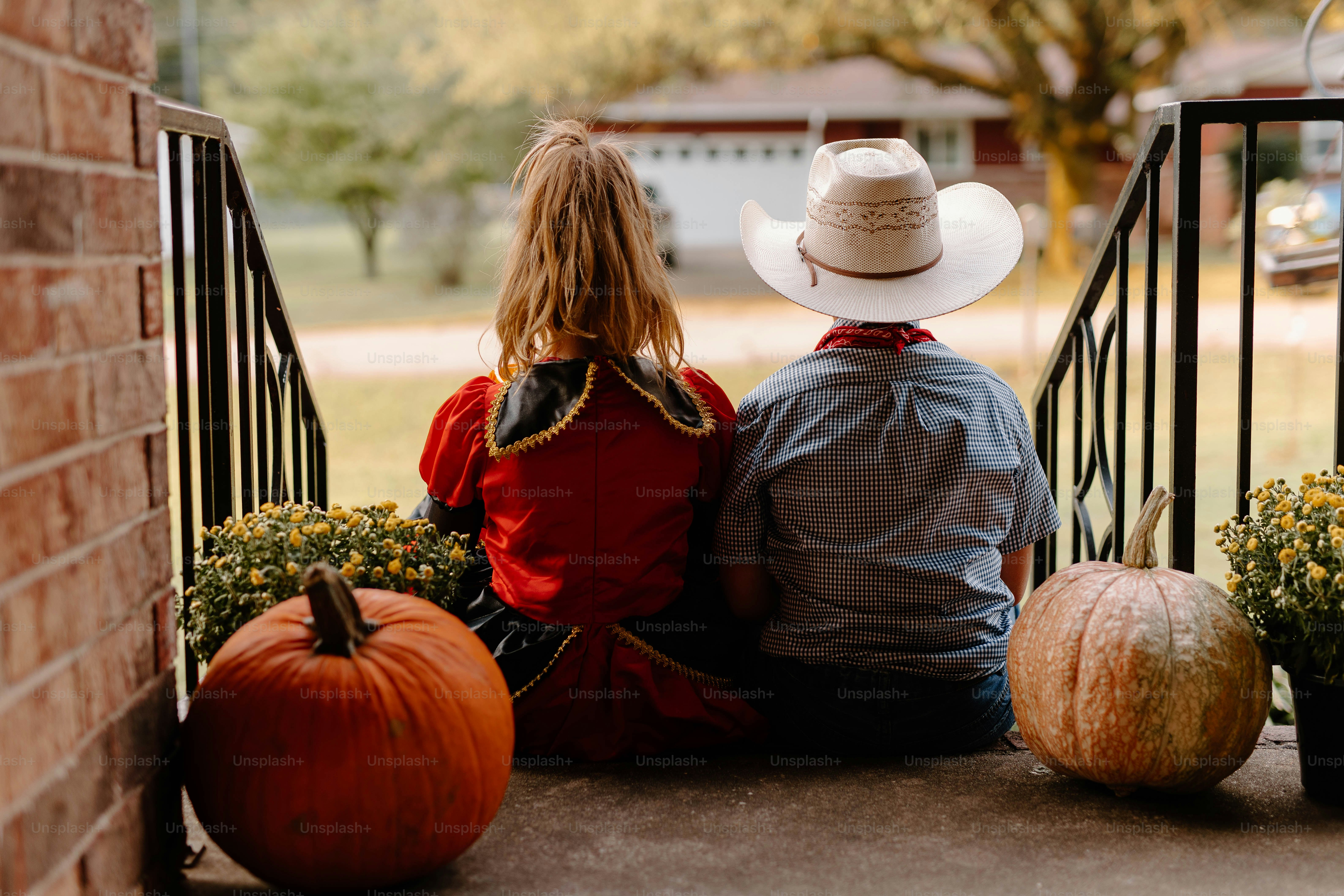 Two children in costumes sit on porch steps with pumpkins.