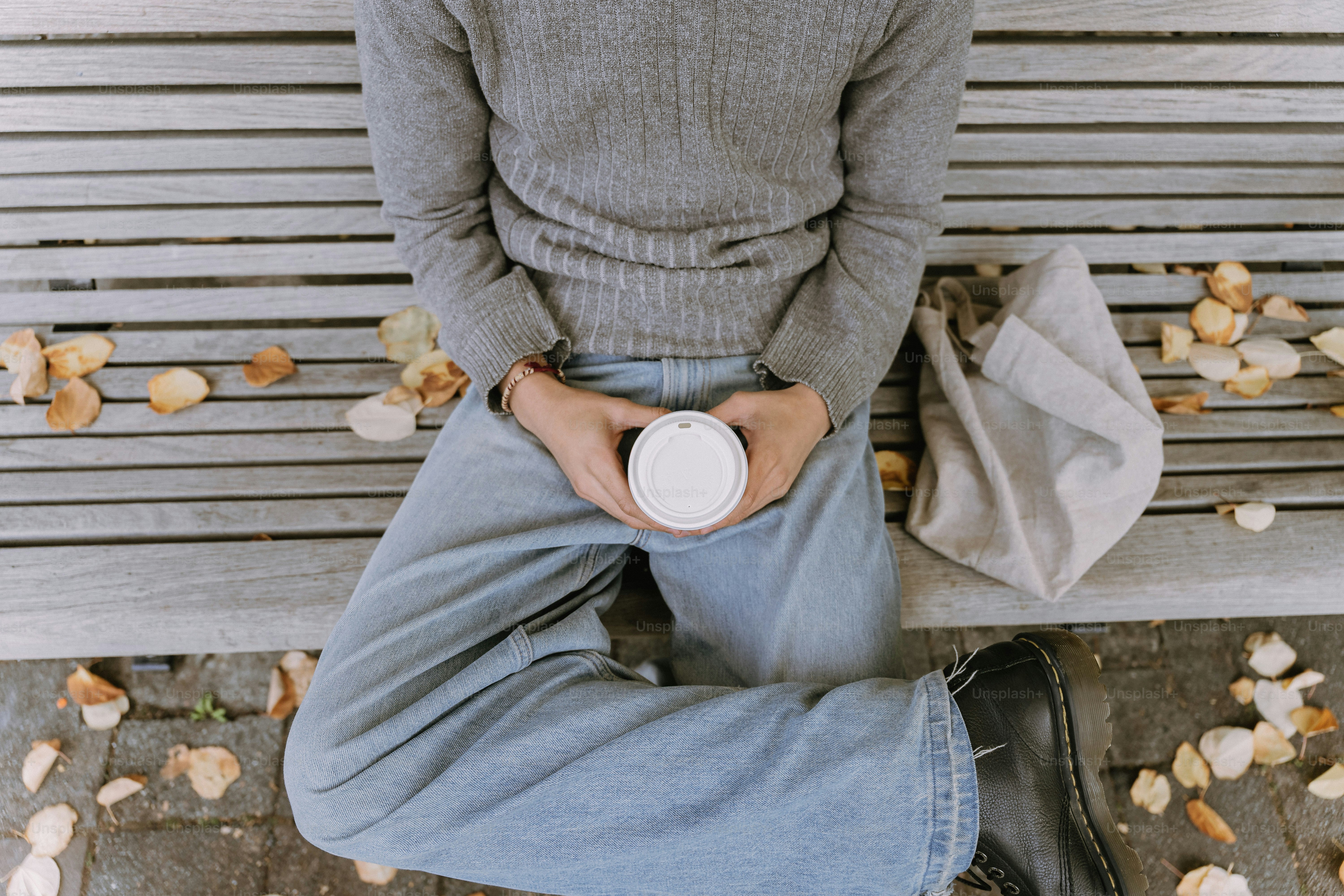 Person holding a coffee cup on a bench with leaves