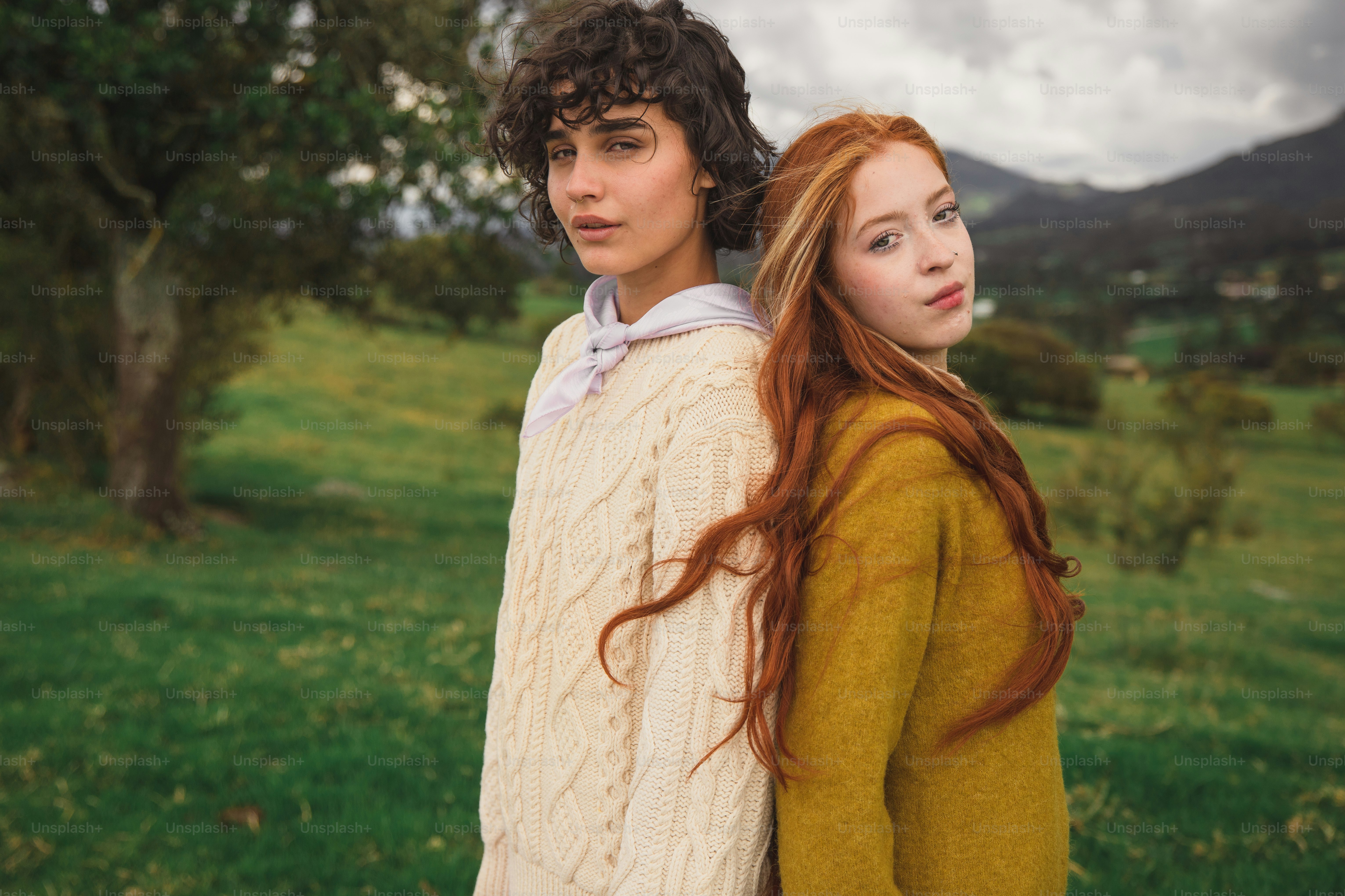 Two young women stand back to back outdoors.