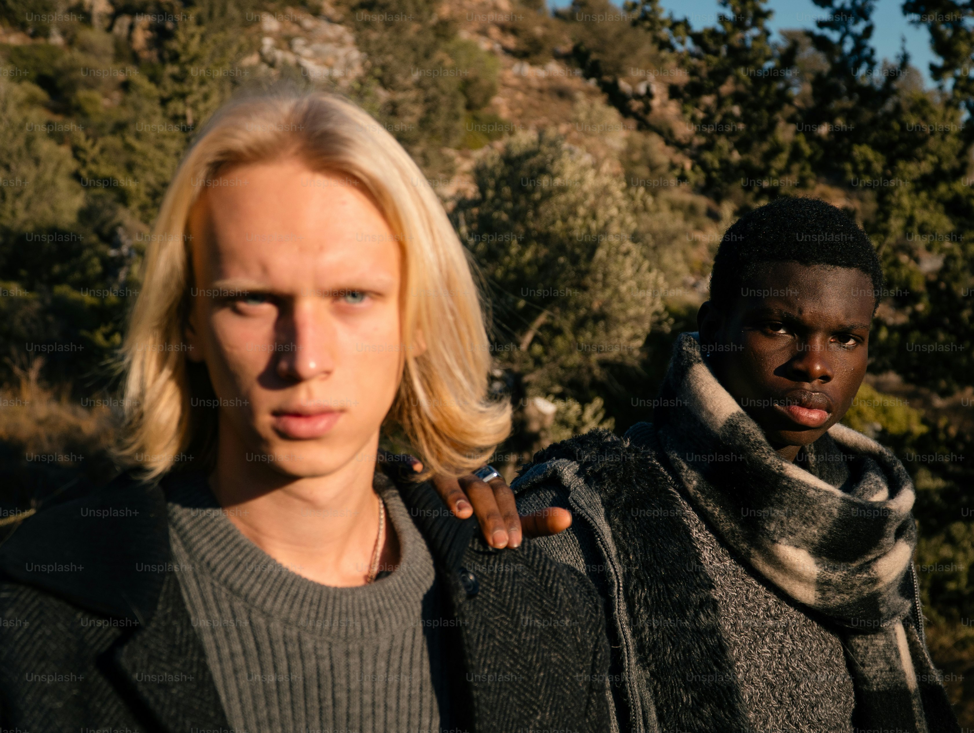 Two young men stand outdoors with trees and hills.
