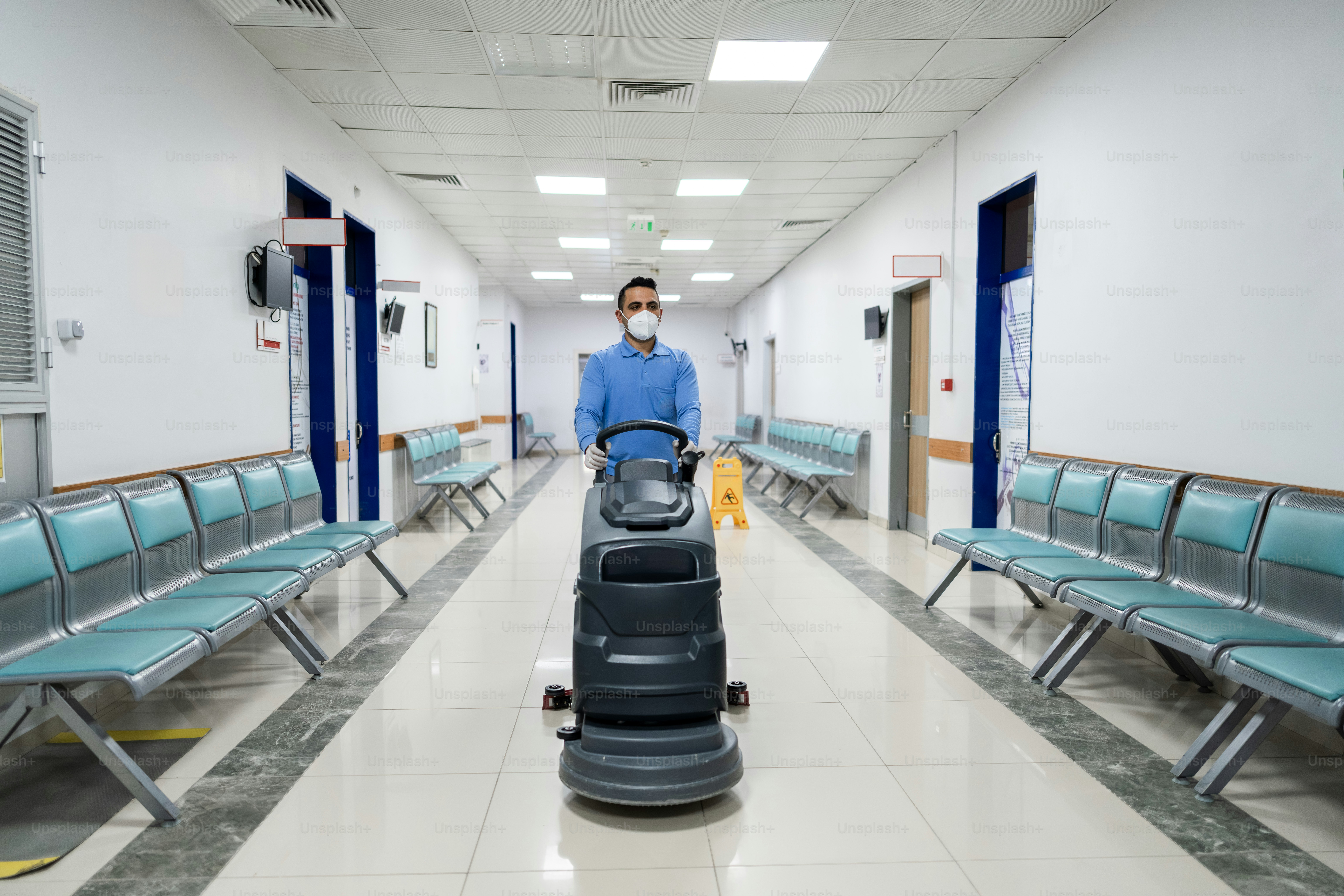 Man with mask cleans hospital hallway with floor polisher.