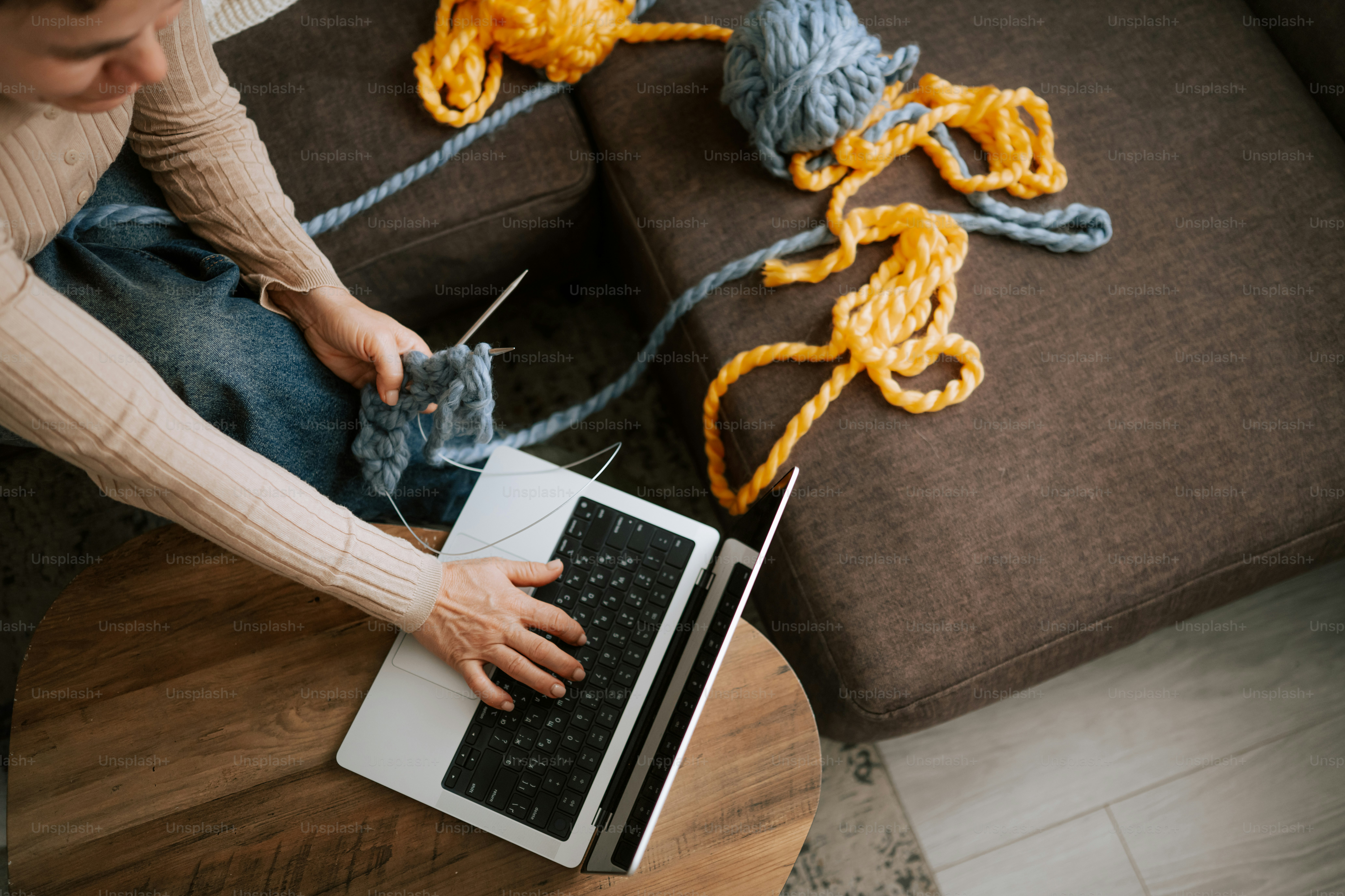 Mulher tricotando enquanto usa um laptop no sofá.