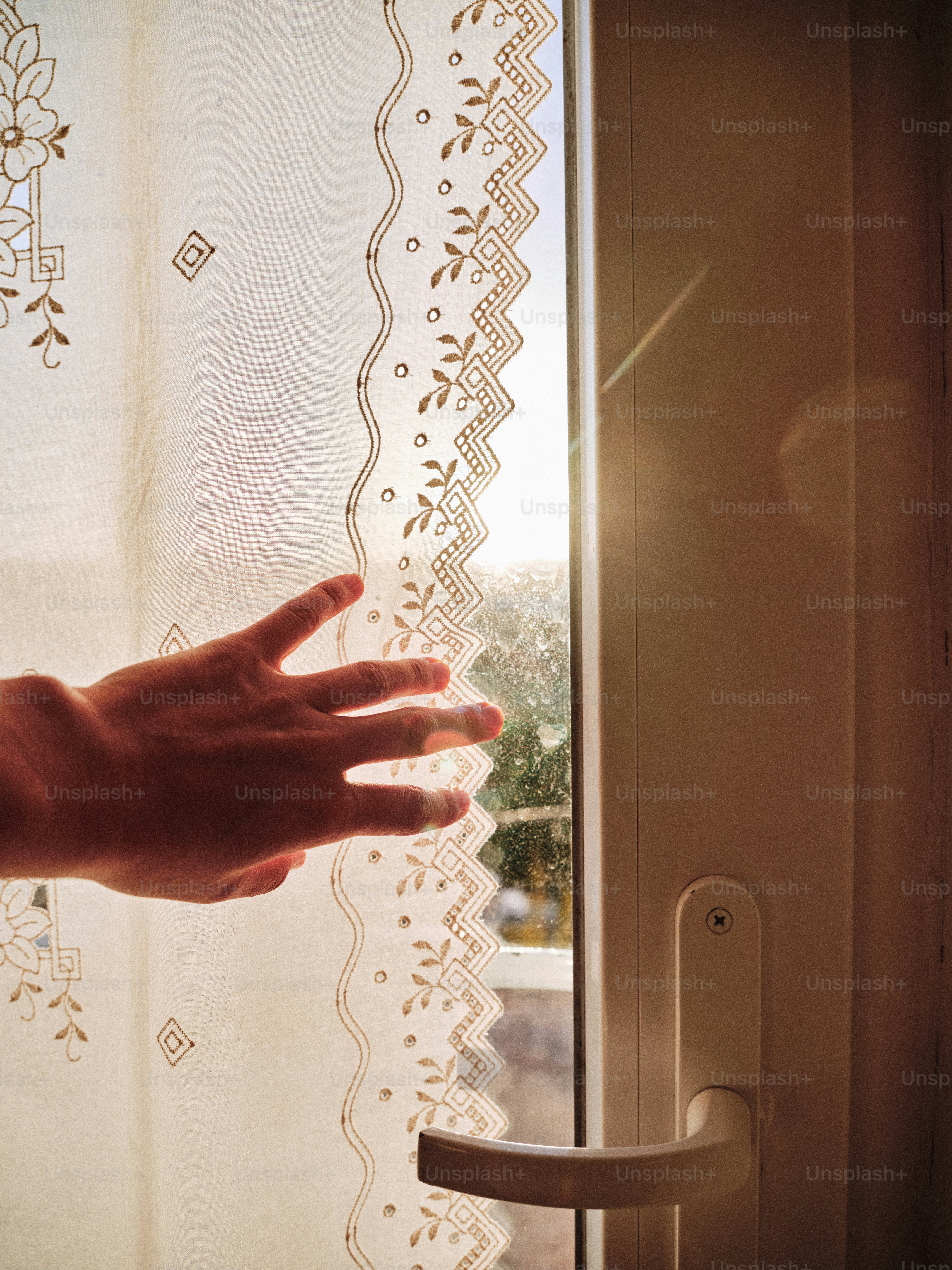 Mano extendida hacia una ventana iluminada por el sol con cortina transparente.