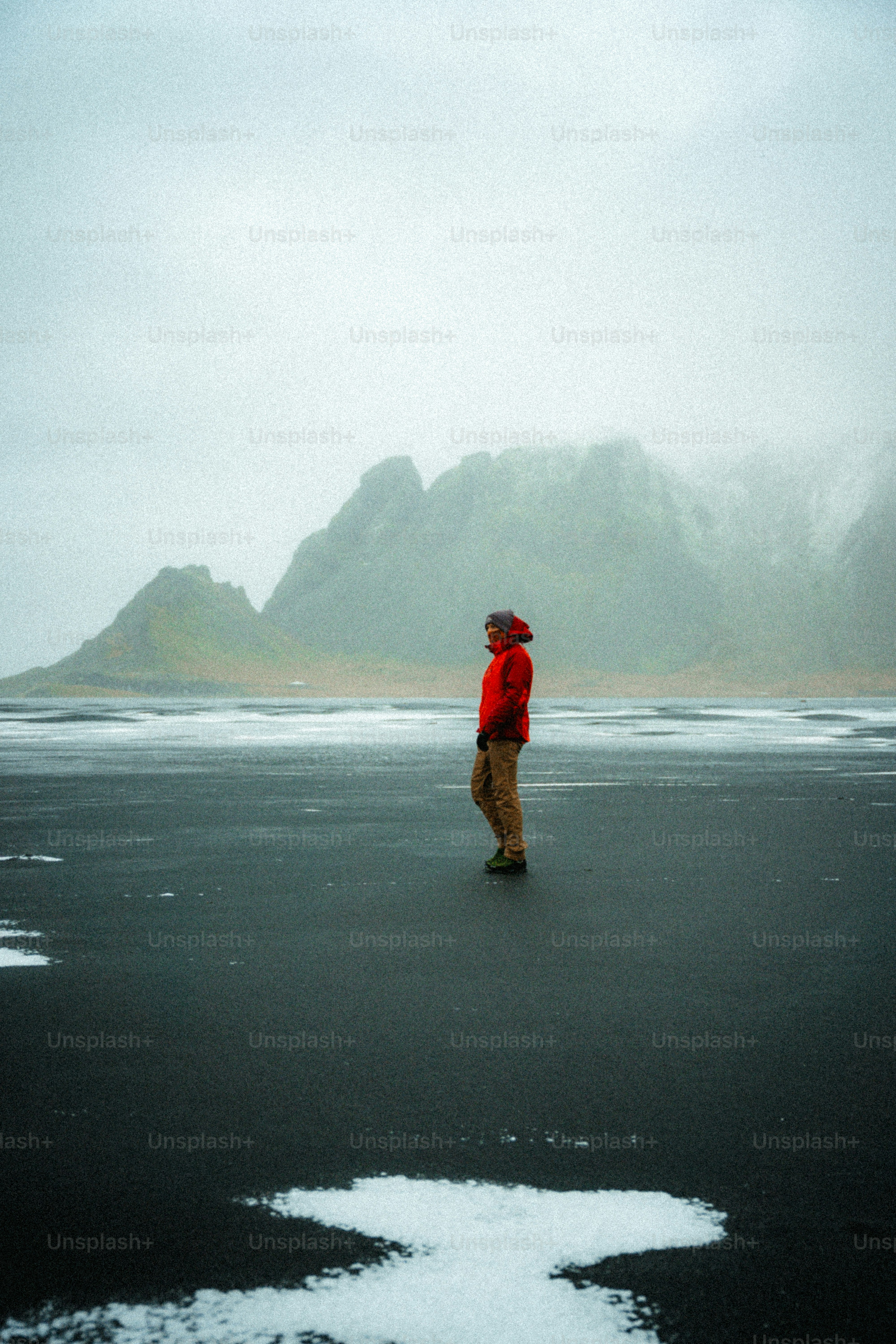 Person in red jacket on black sand beach with mountains.