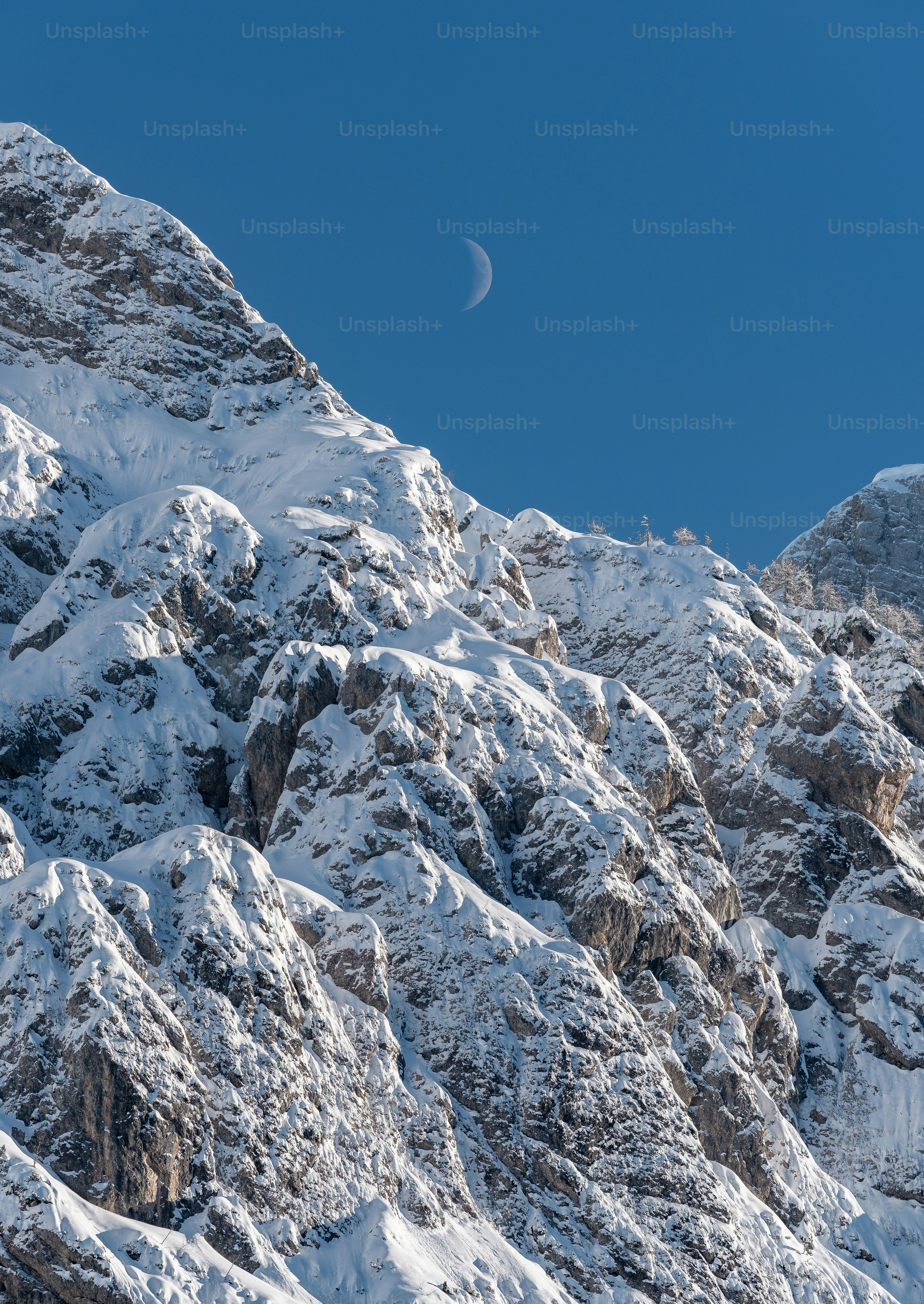 Crescent moon over snow-covered mountains under a clear blue sky.