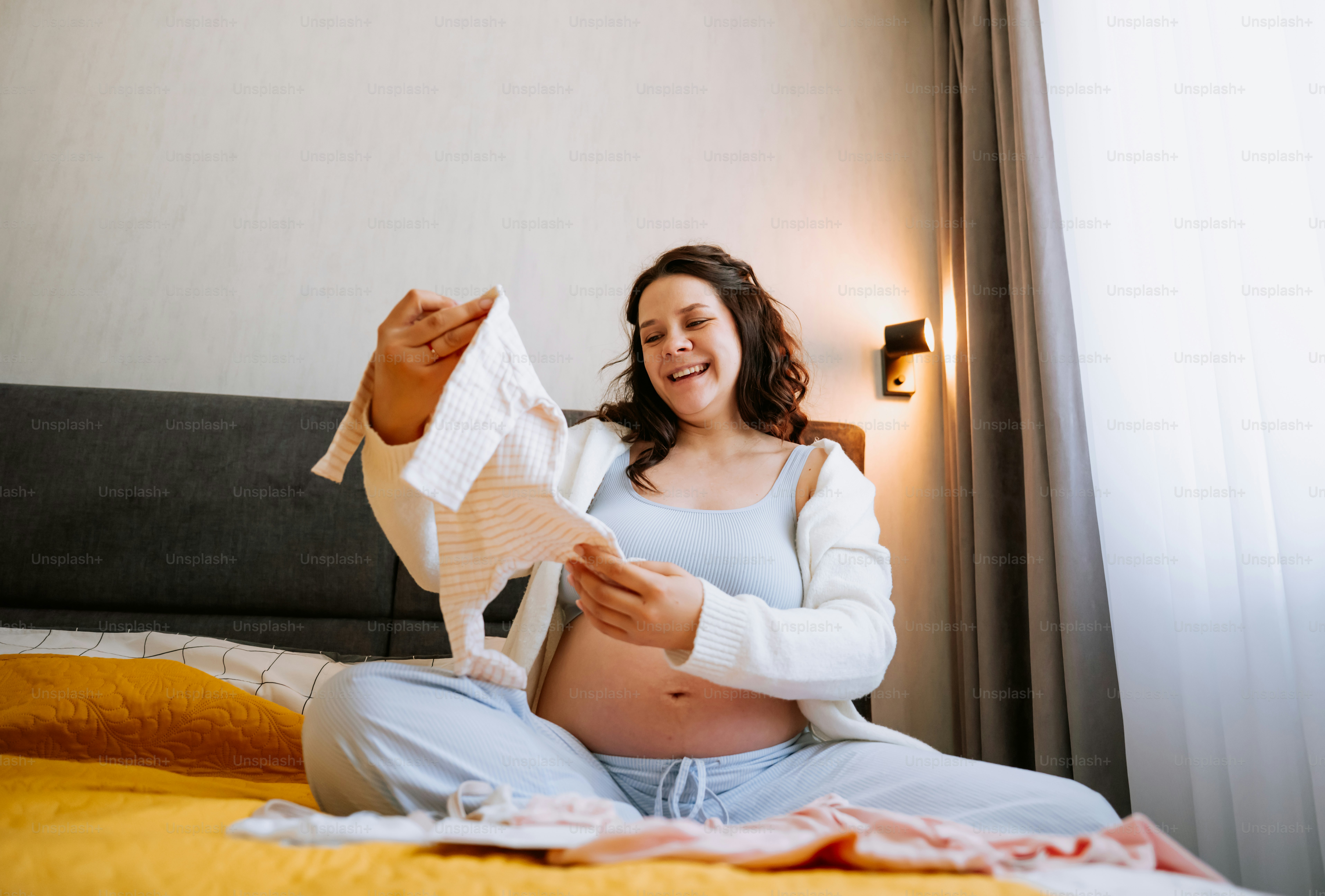 Pregnant woman holding baby clothes on bed