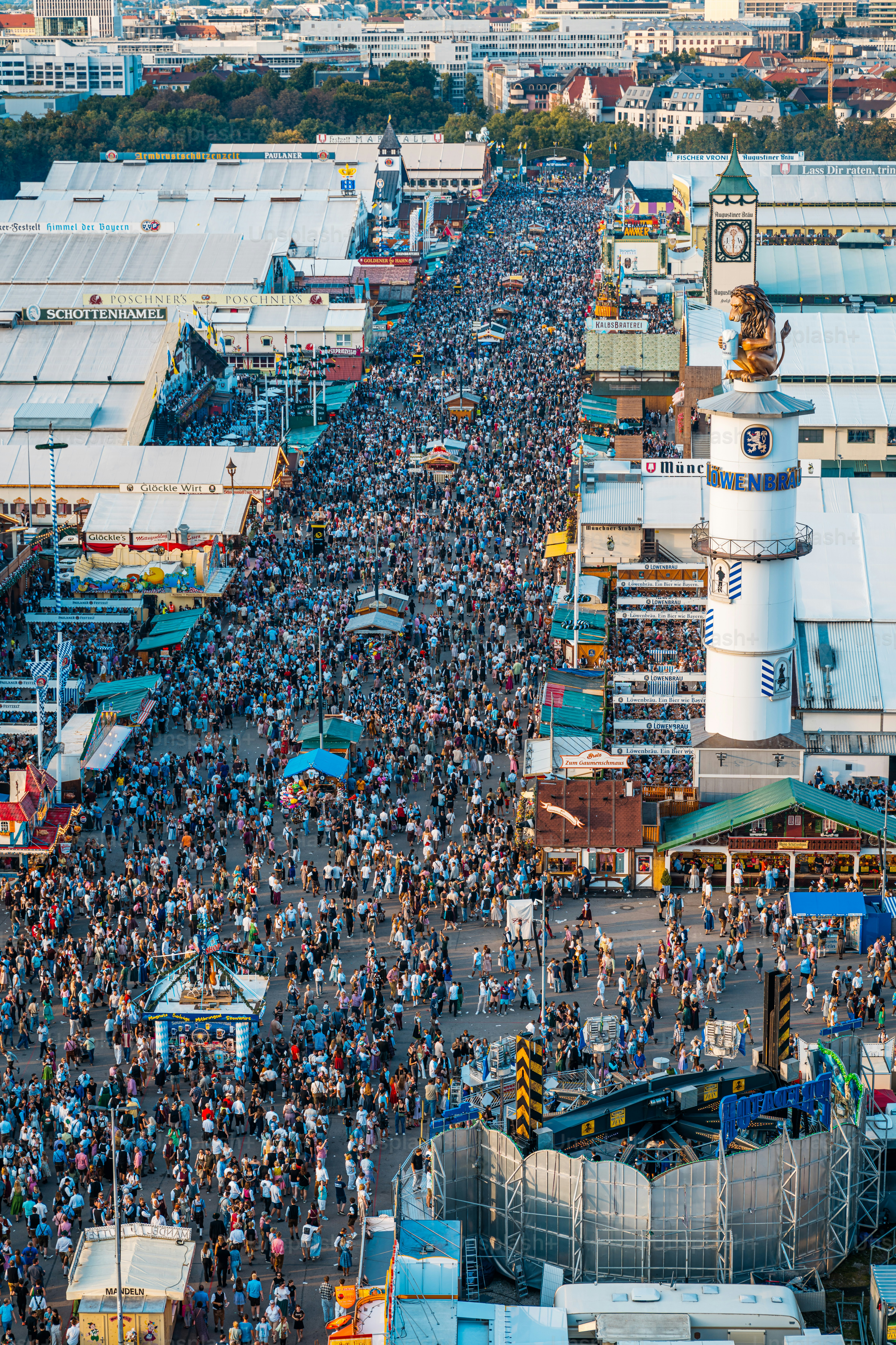 Eine sonnenbeschienene, geschäftige Münchner Straße beim weltberühmten Volksfest mit dichten Menschenmengen, Bierzelten, Fahrgeschäften und einem hohen Uhrturm, die die traditionelle bayerische Festenergie und -größe verkörpert.