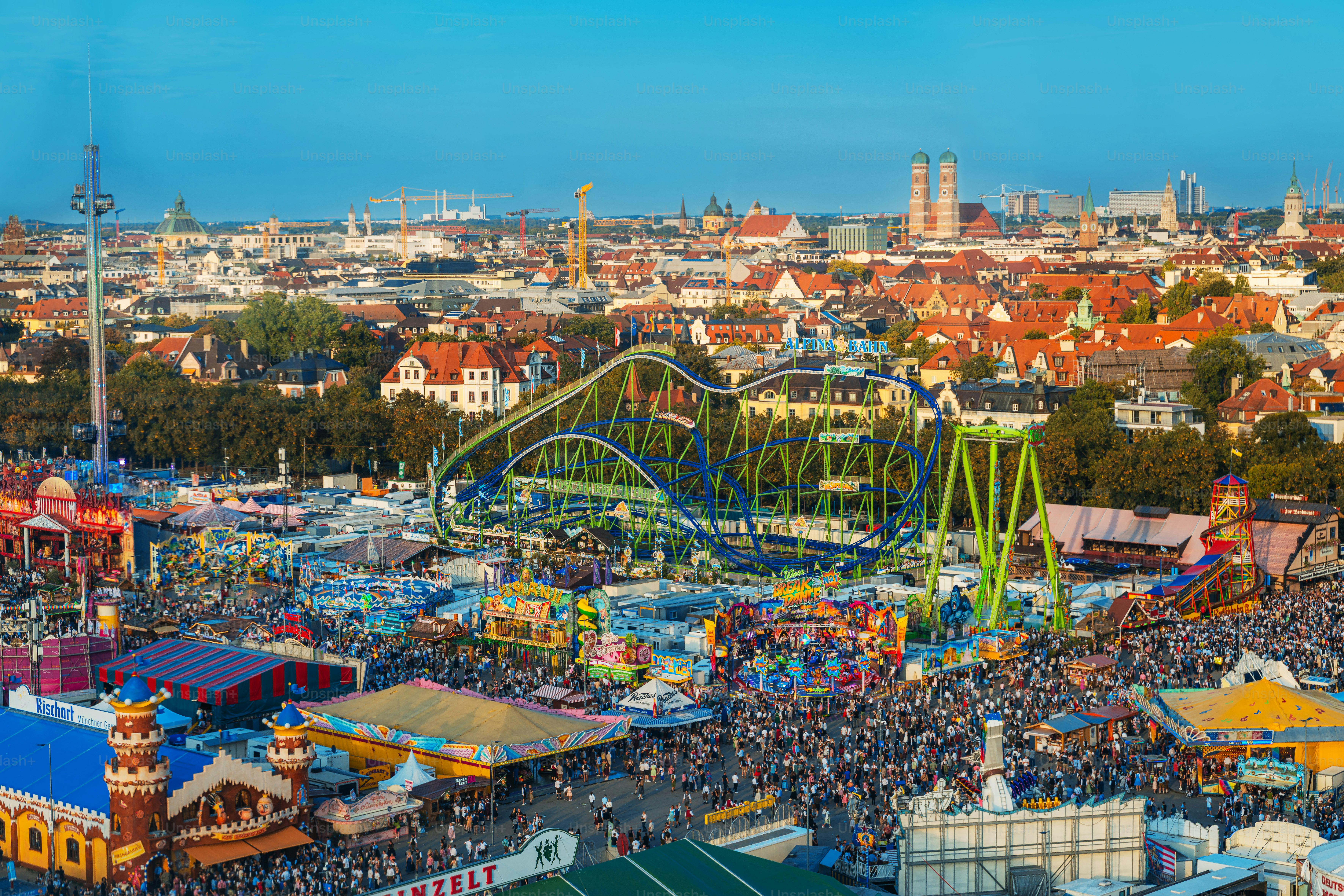 Ein belebter Münchner Jahrmarktsplatz strotzt vor bunten Fahrgeschäften, Essensstanden und jubelnden Menschenmengen während des größten Bierfestivals der Welt. Heller Himmel, festliche Energie und architektonische Stadtlandschaften schaffen eine dynamische Feier.