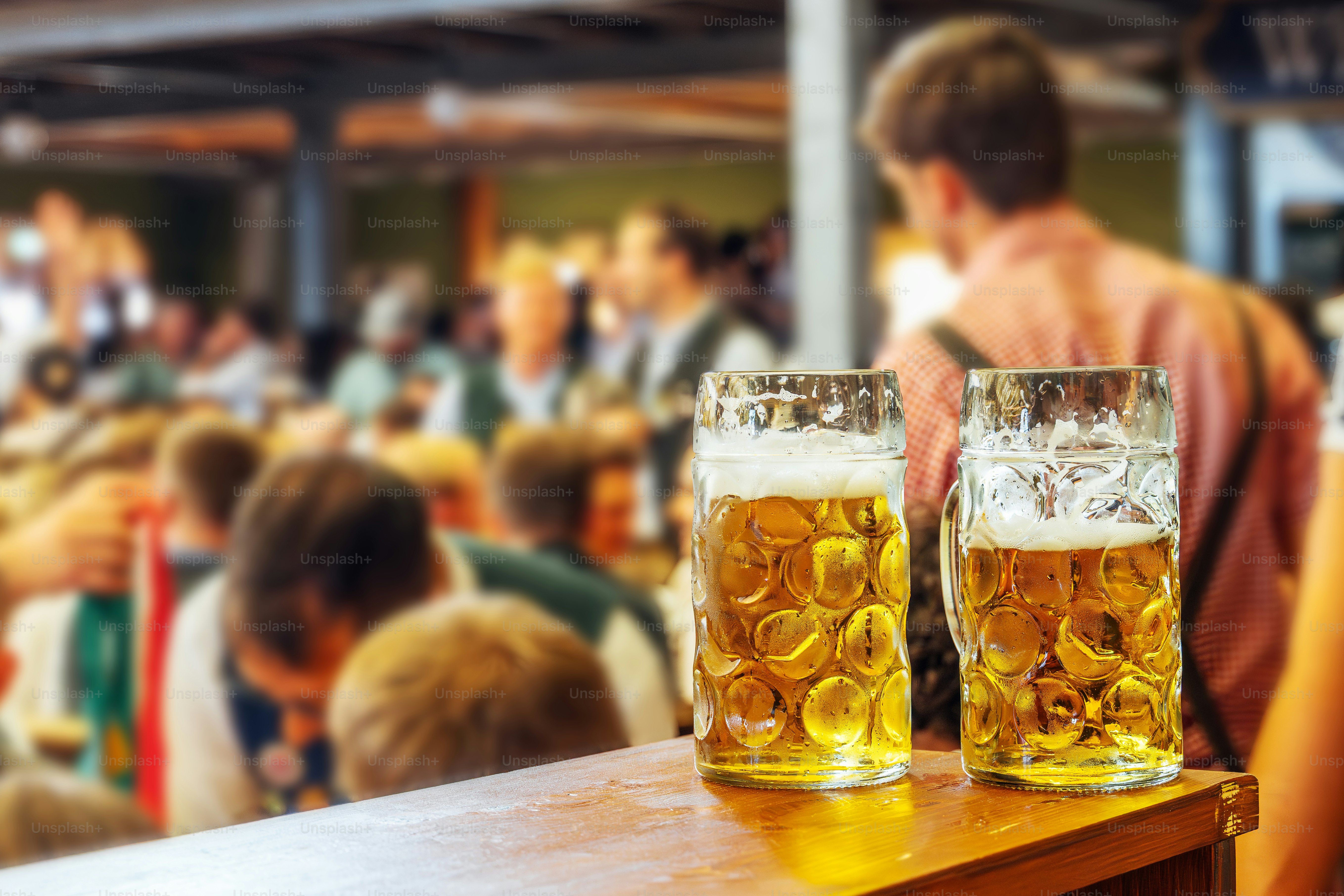 Two glass mugs on a wooden bar overlook a bustling crowd at Munich's Wiesn, capturing the festive mood, camaraderie, and beer-loving spirit of the world’s largest beer festival.