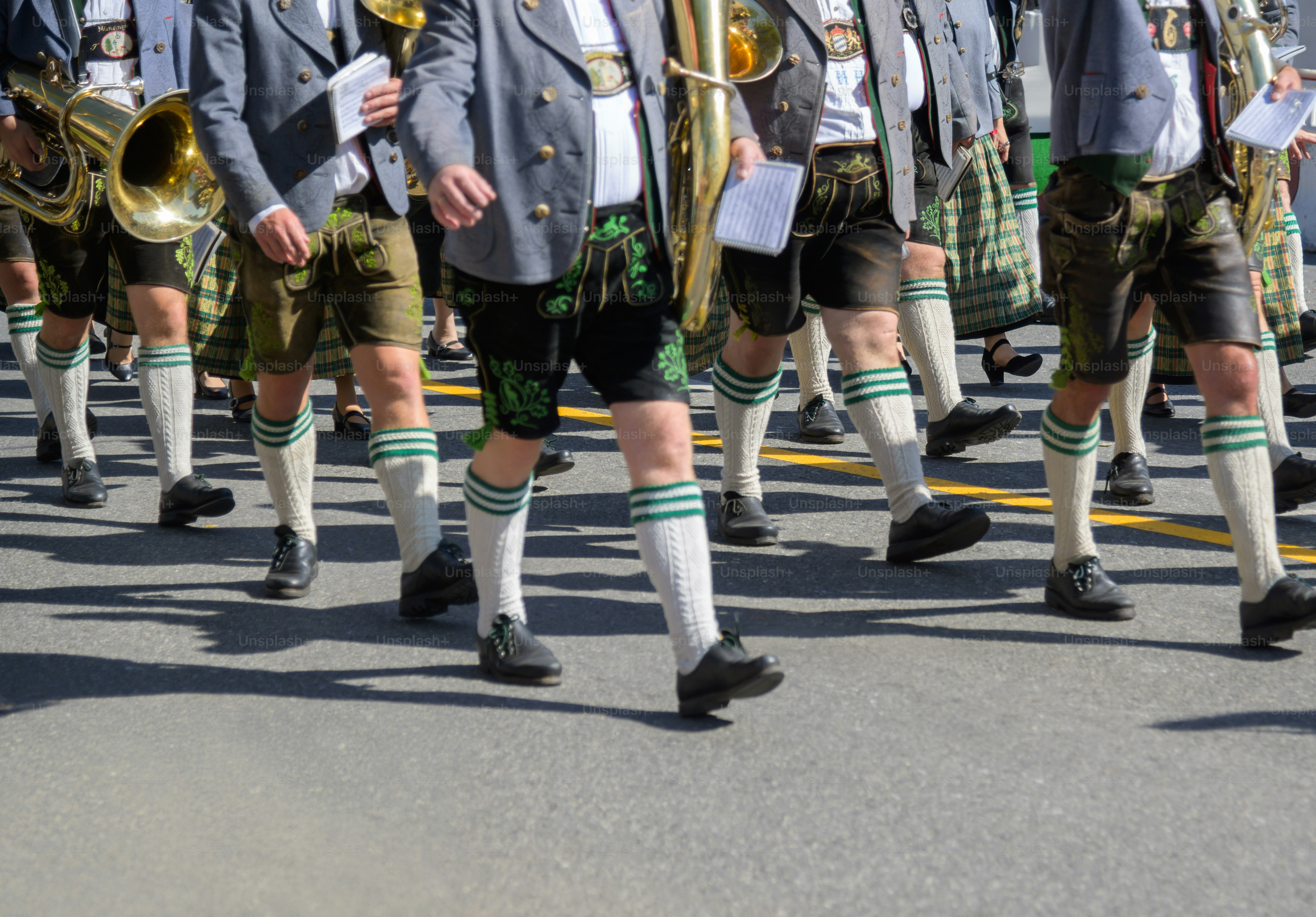 A lively street parade featuring men in lederhosen and women in dirndls, playing brass instruments as they march down a sunlit road. Festive, cultural atmosphere with traditional attire and music.