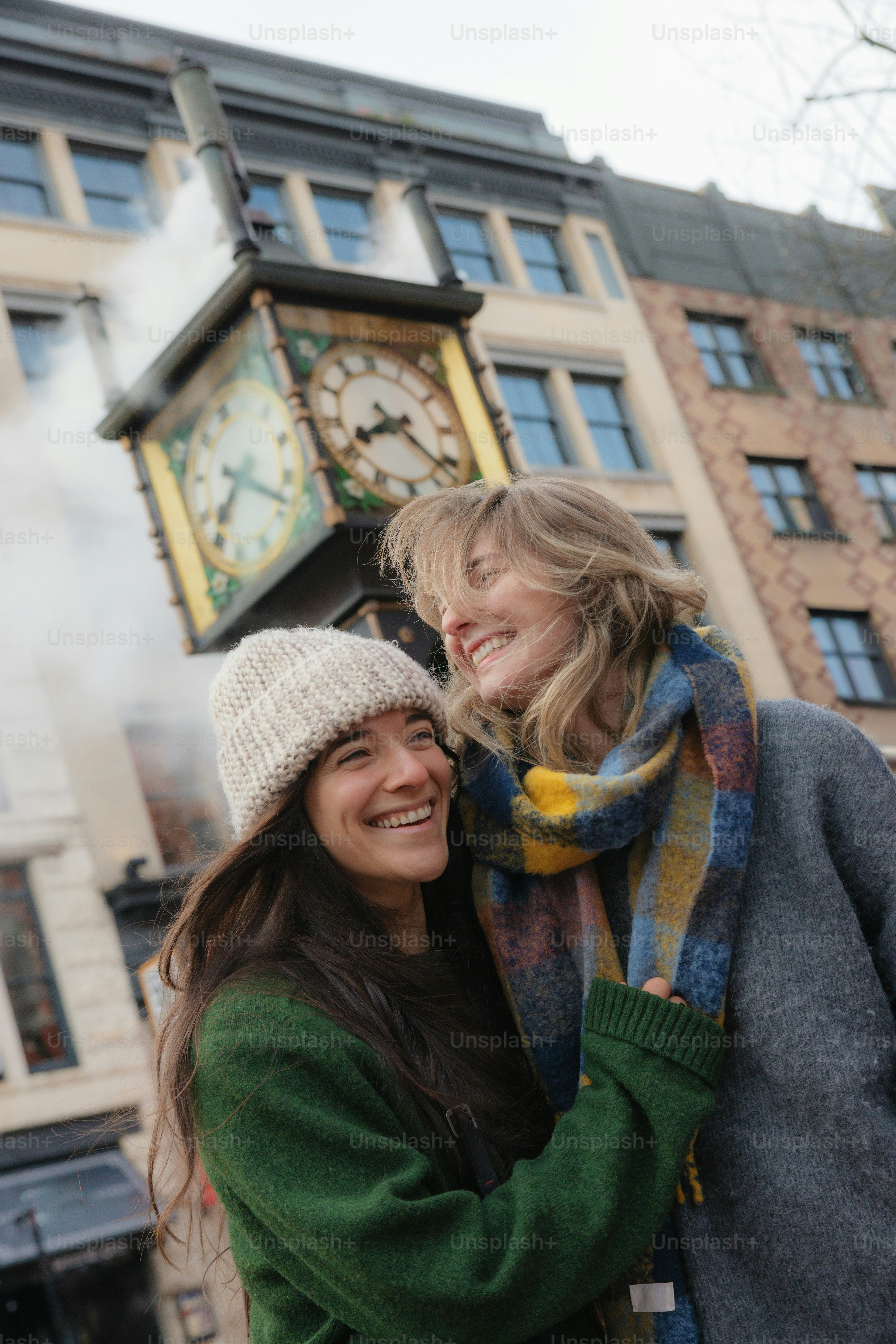Two smiling women embrace near a clock tower.