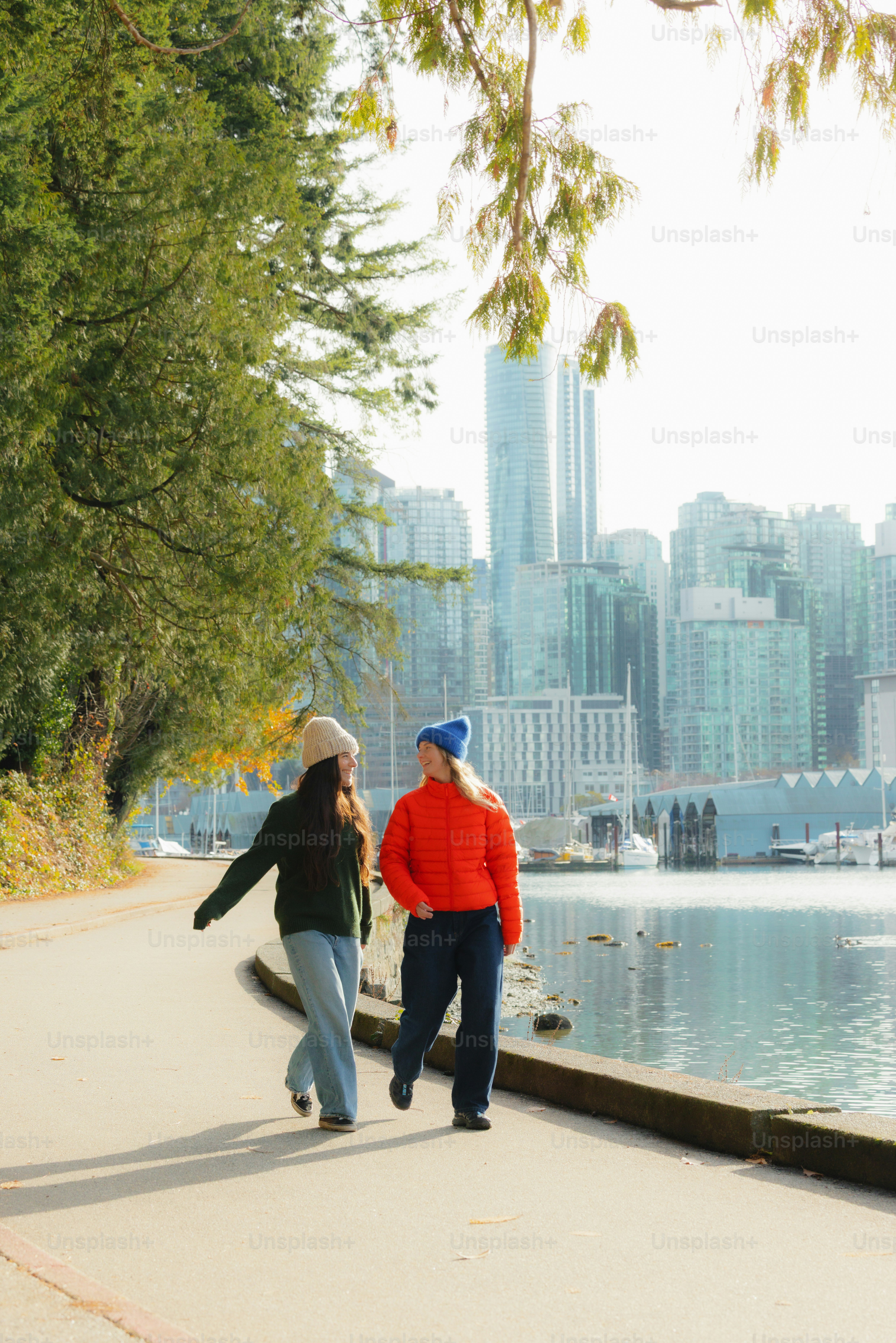 Two women walk along a waterfront path with city skyline.