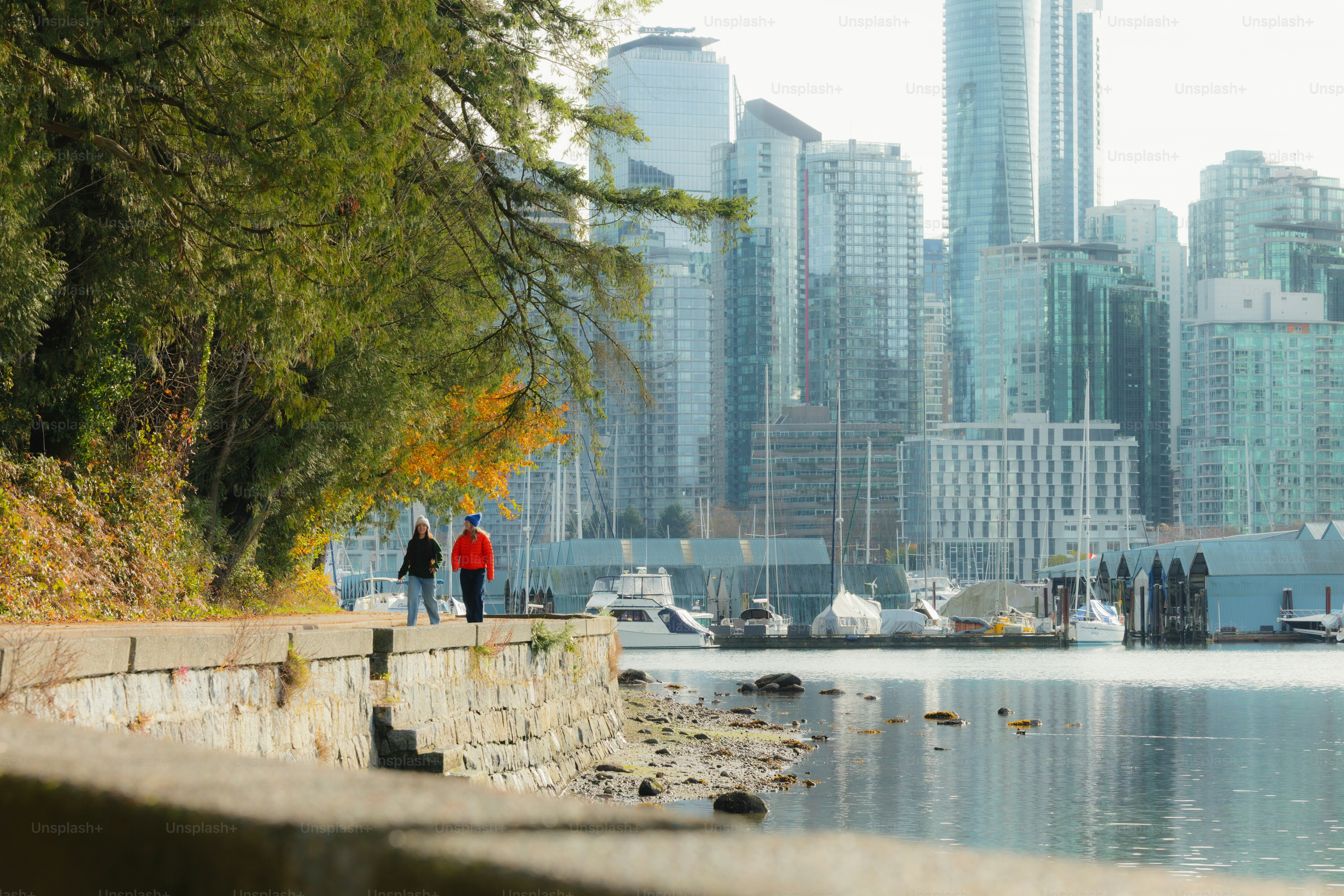 Two people walk along a waterfront path with city skyline.