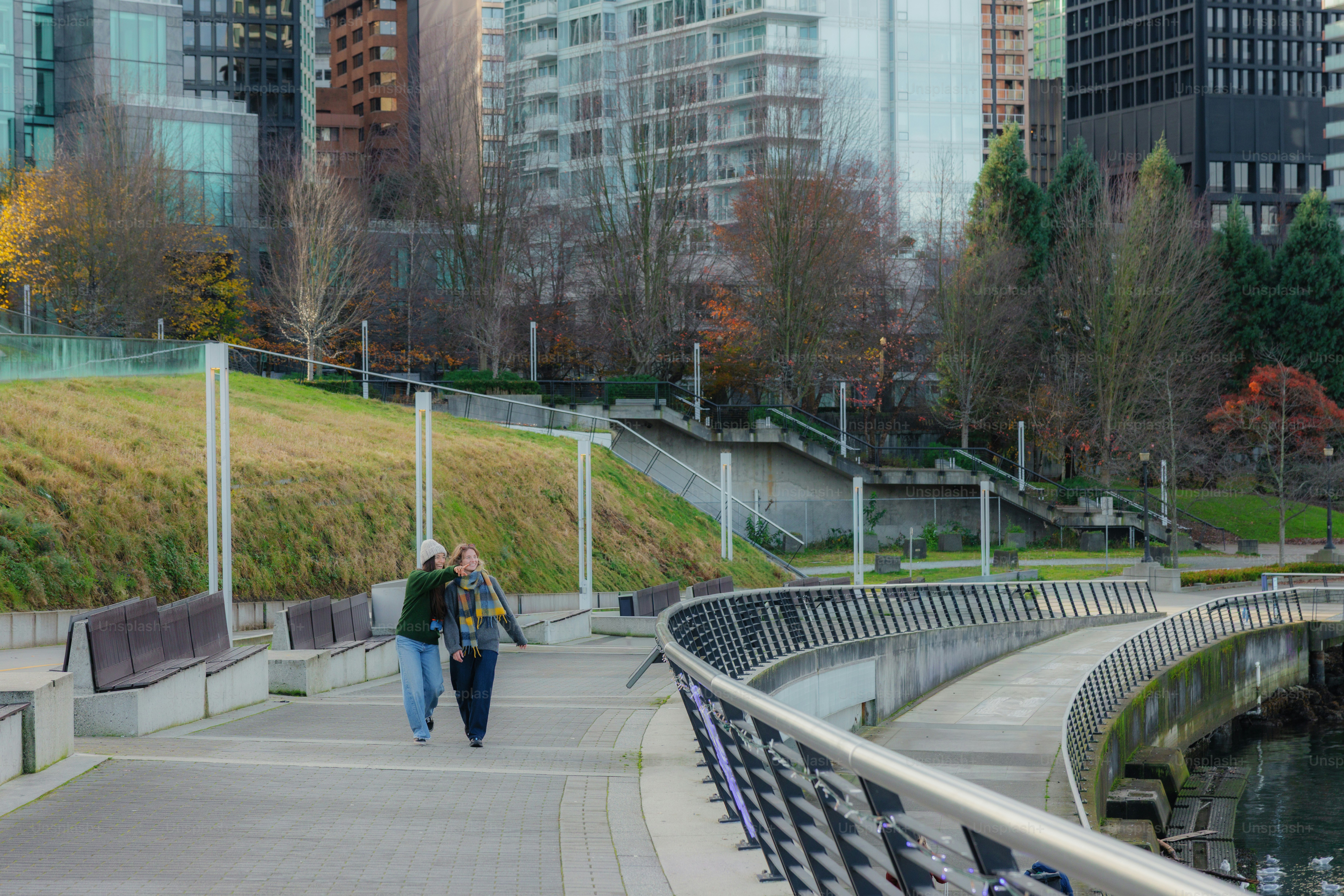 Two people walk along a waterfront path with city buildings.