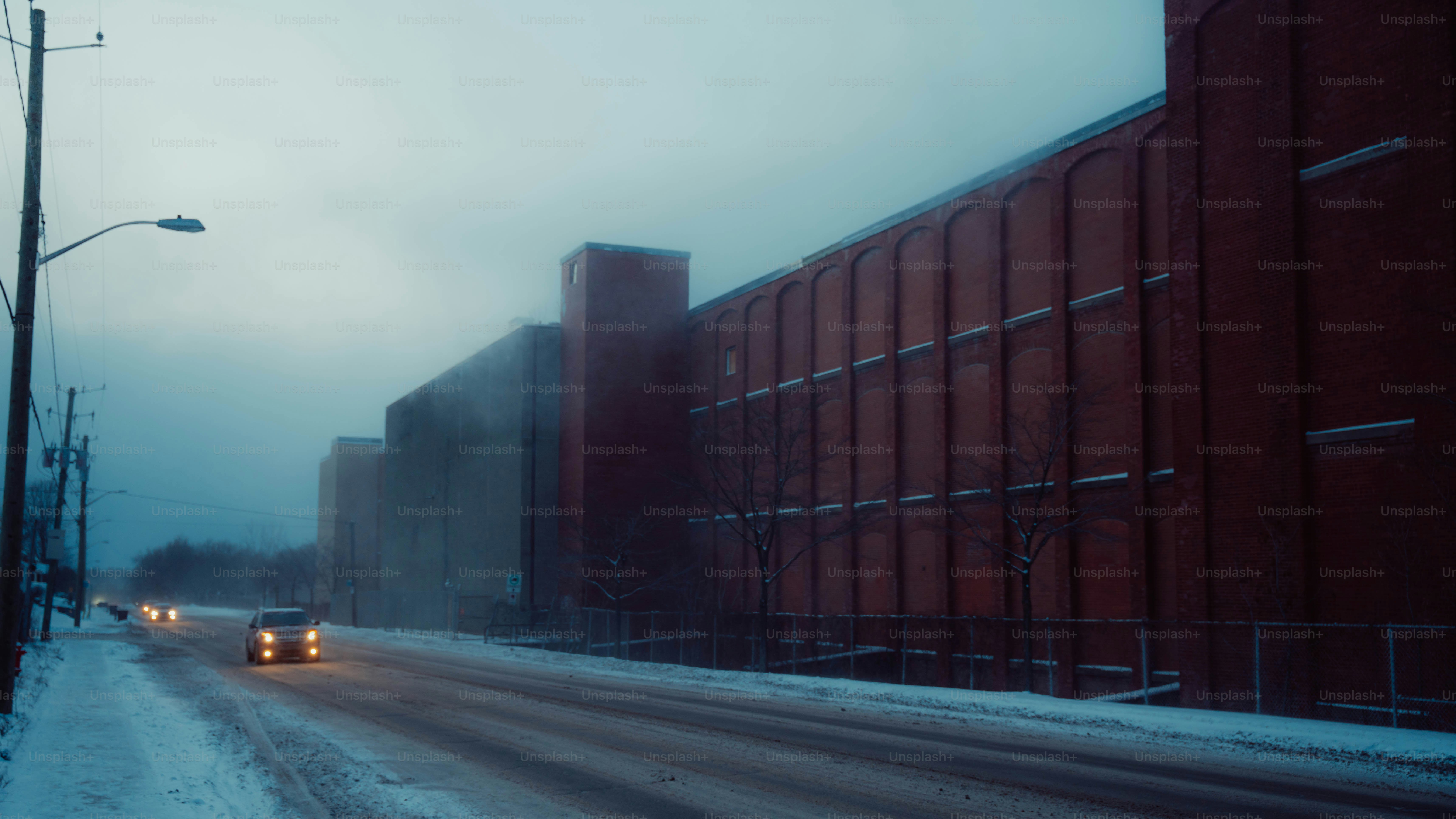 Cars drive past a large red brick building in snow.