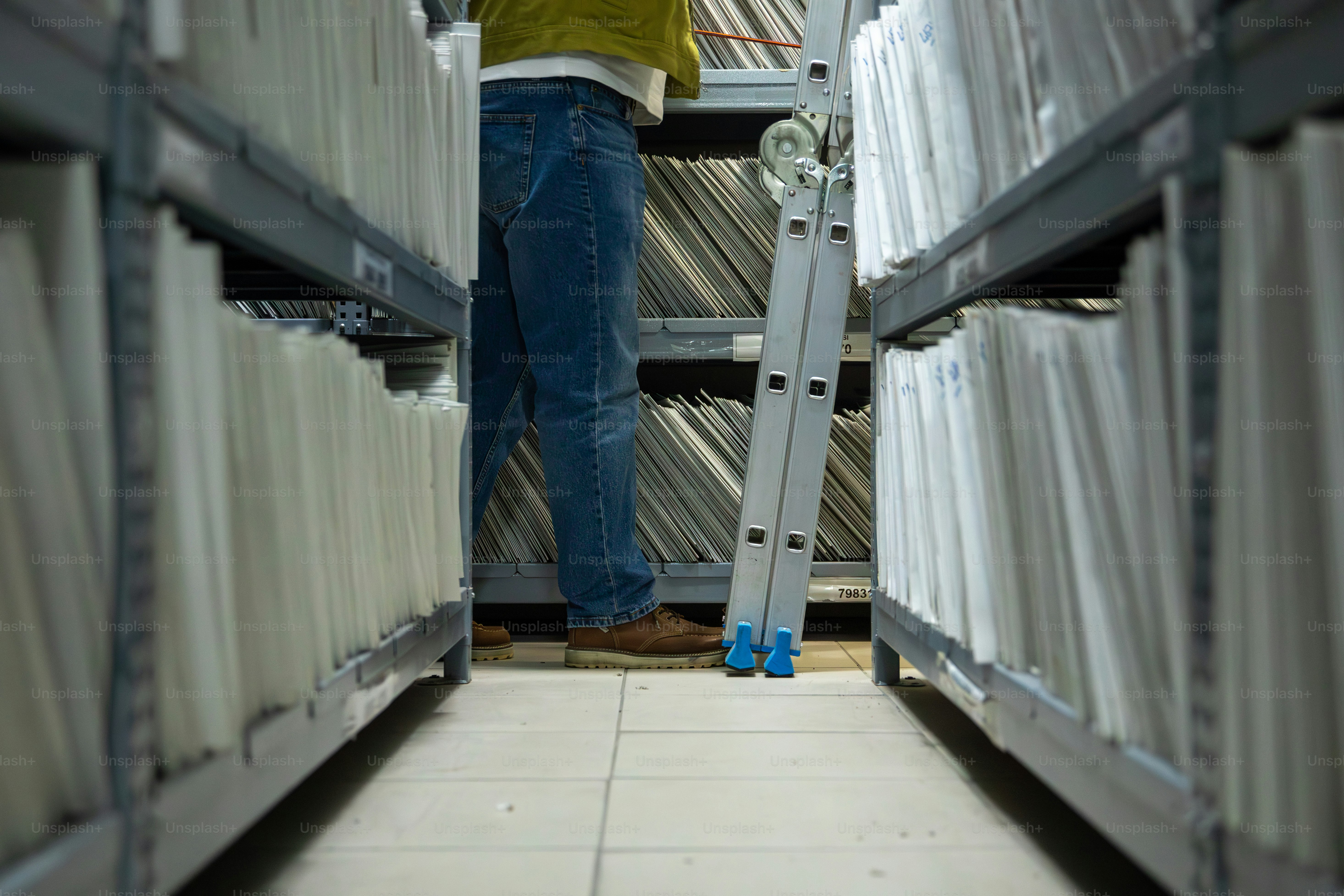 사다리가 있는 높은 선반 위 파일을 집어 들고 있는 사람.