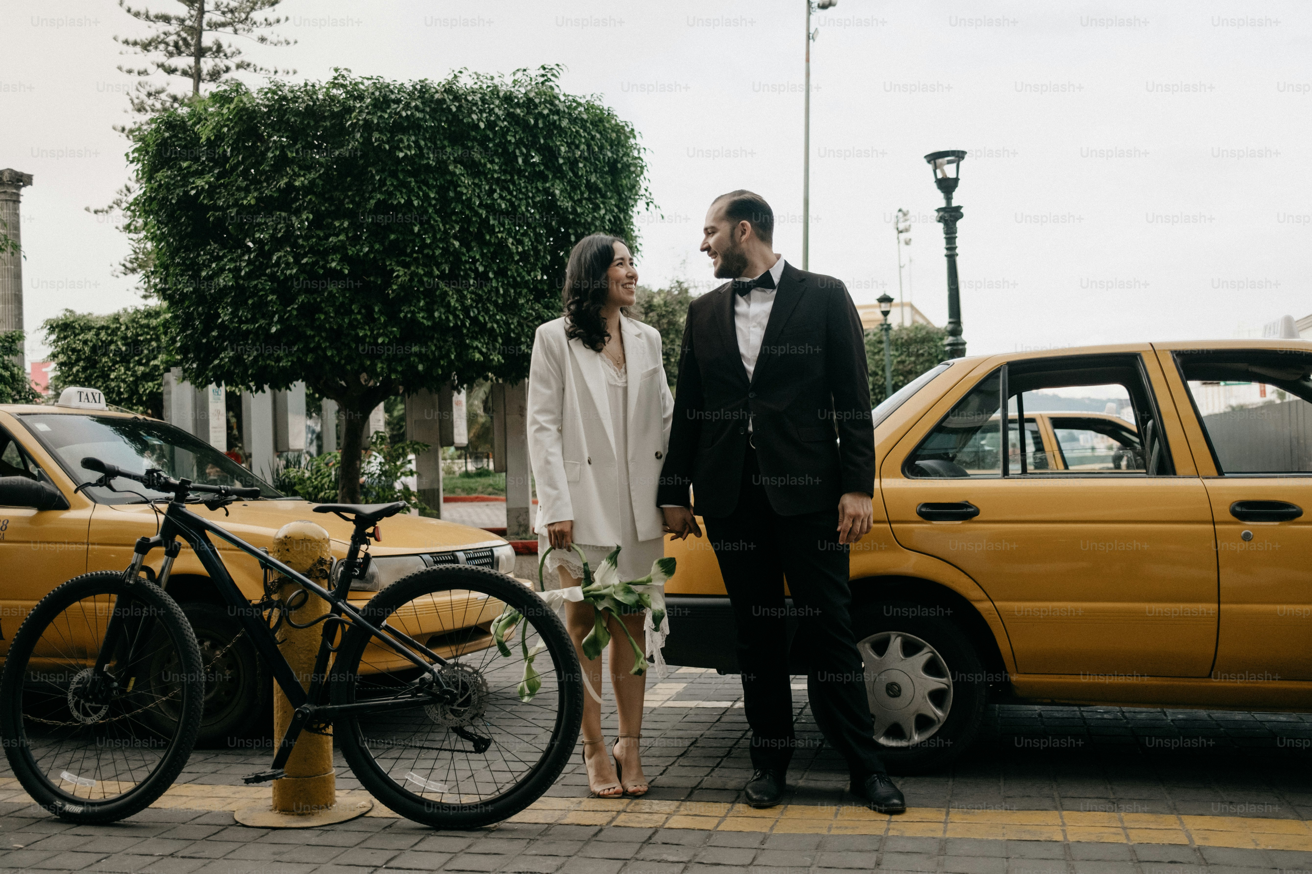 Newlyweds holding hands near yellow taxis and bicycle.
