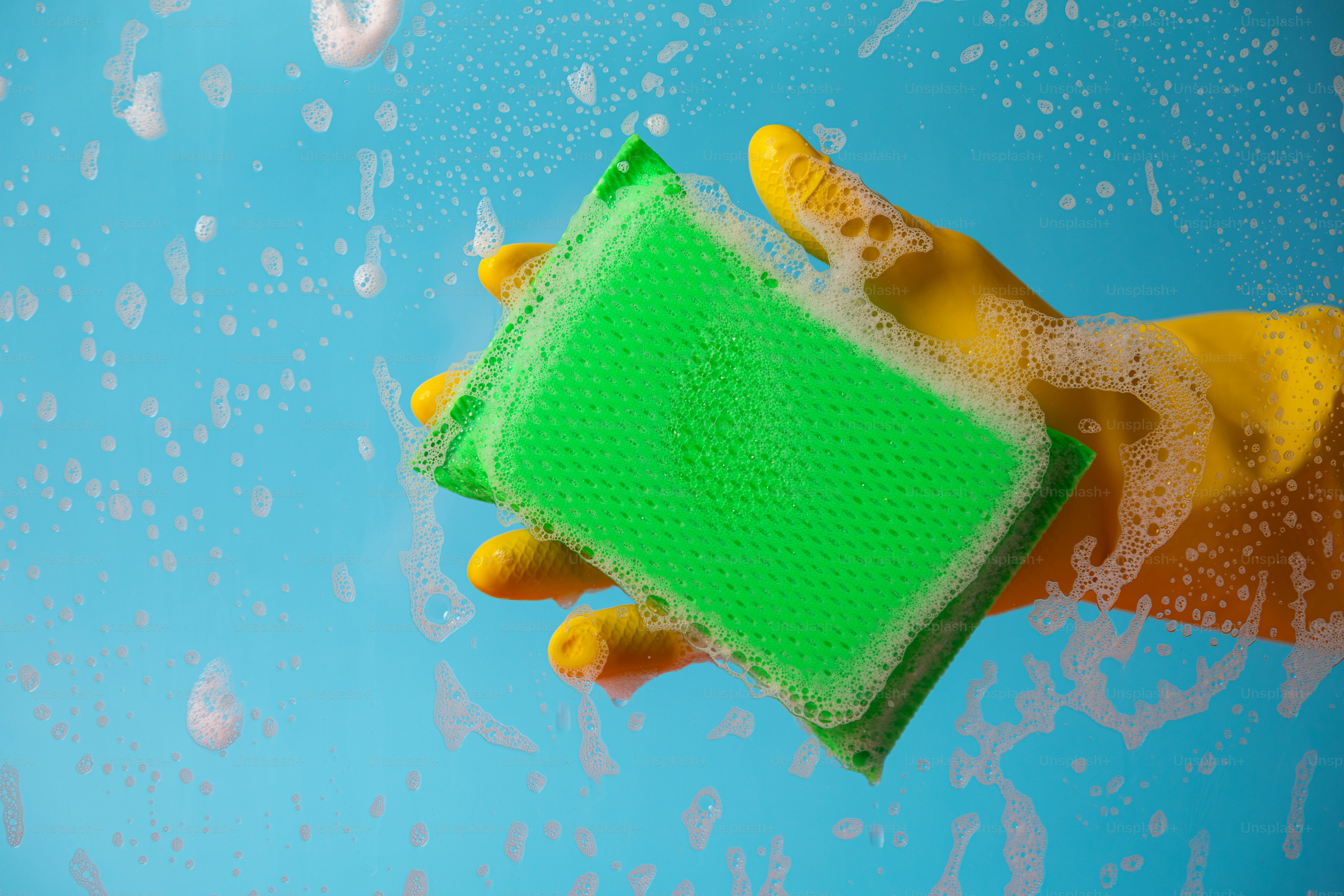 Main dans un gant jaune tenant une éponge verte avec des bulles de savon