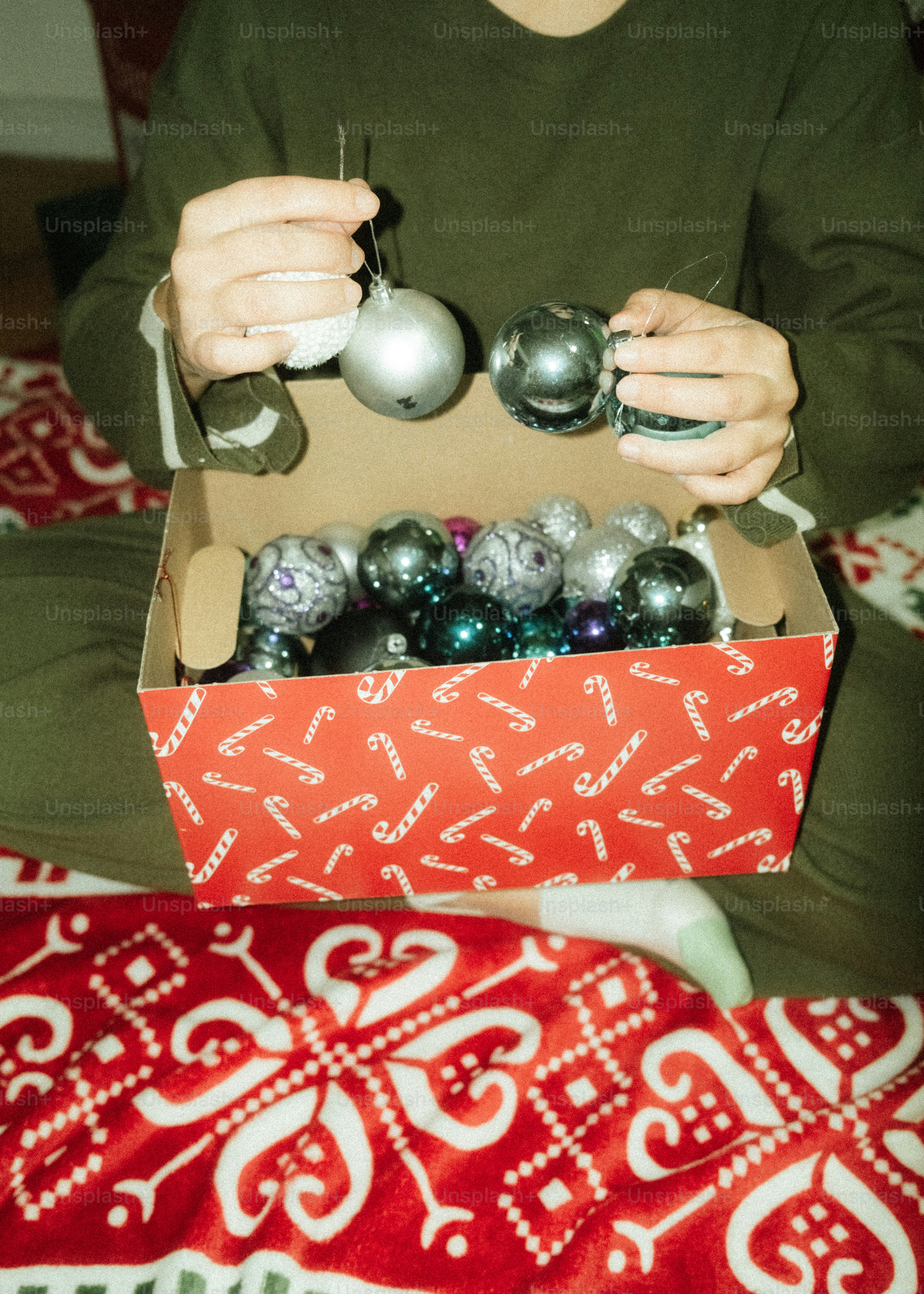 Hands holding christmas ornaments above a box of decorations.