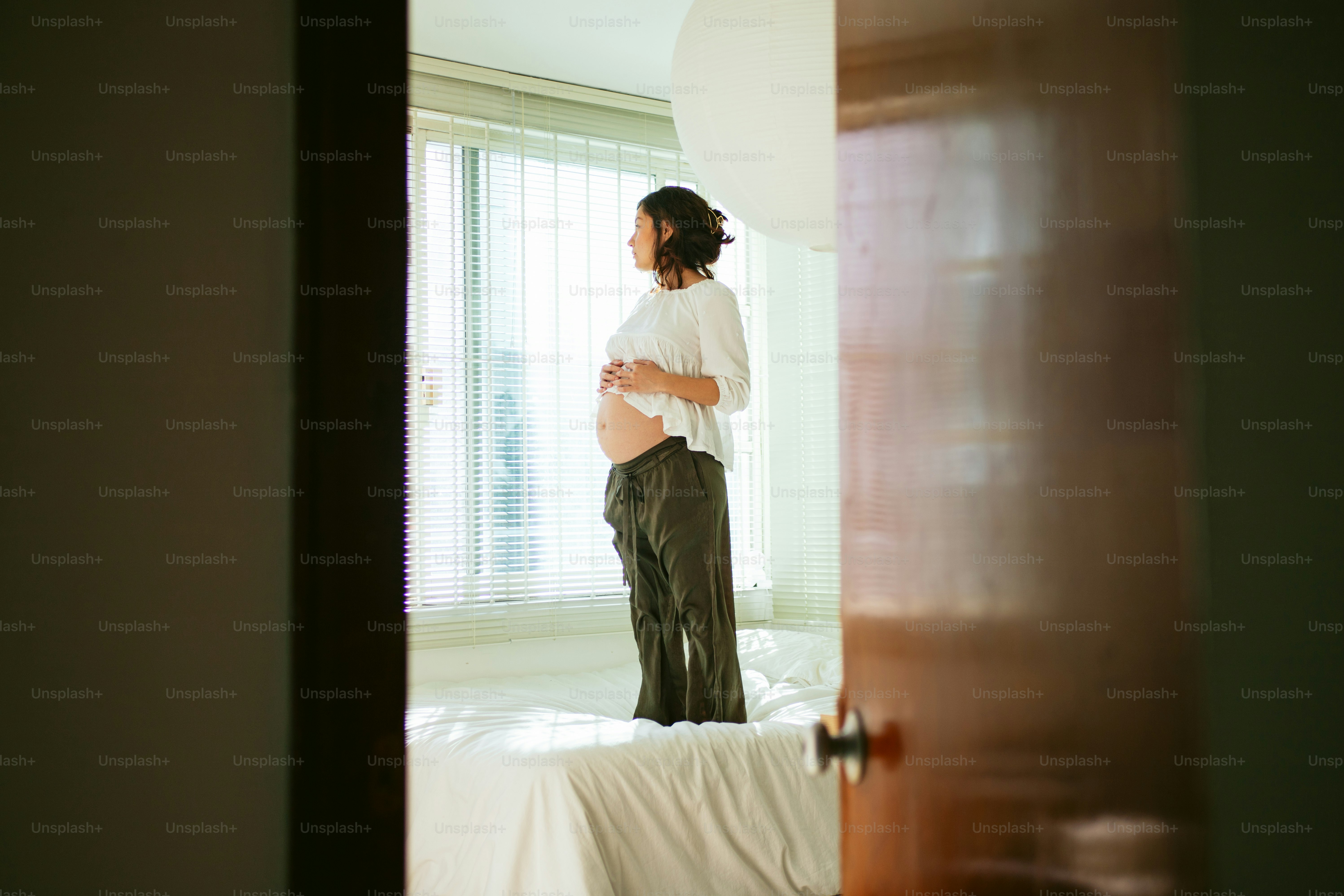 Pregnant woman standing by the window, holding belly.