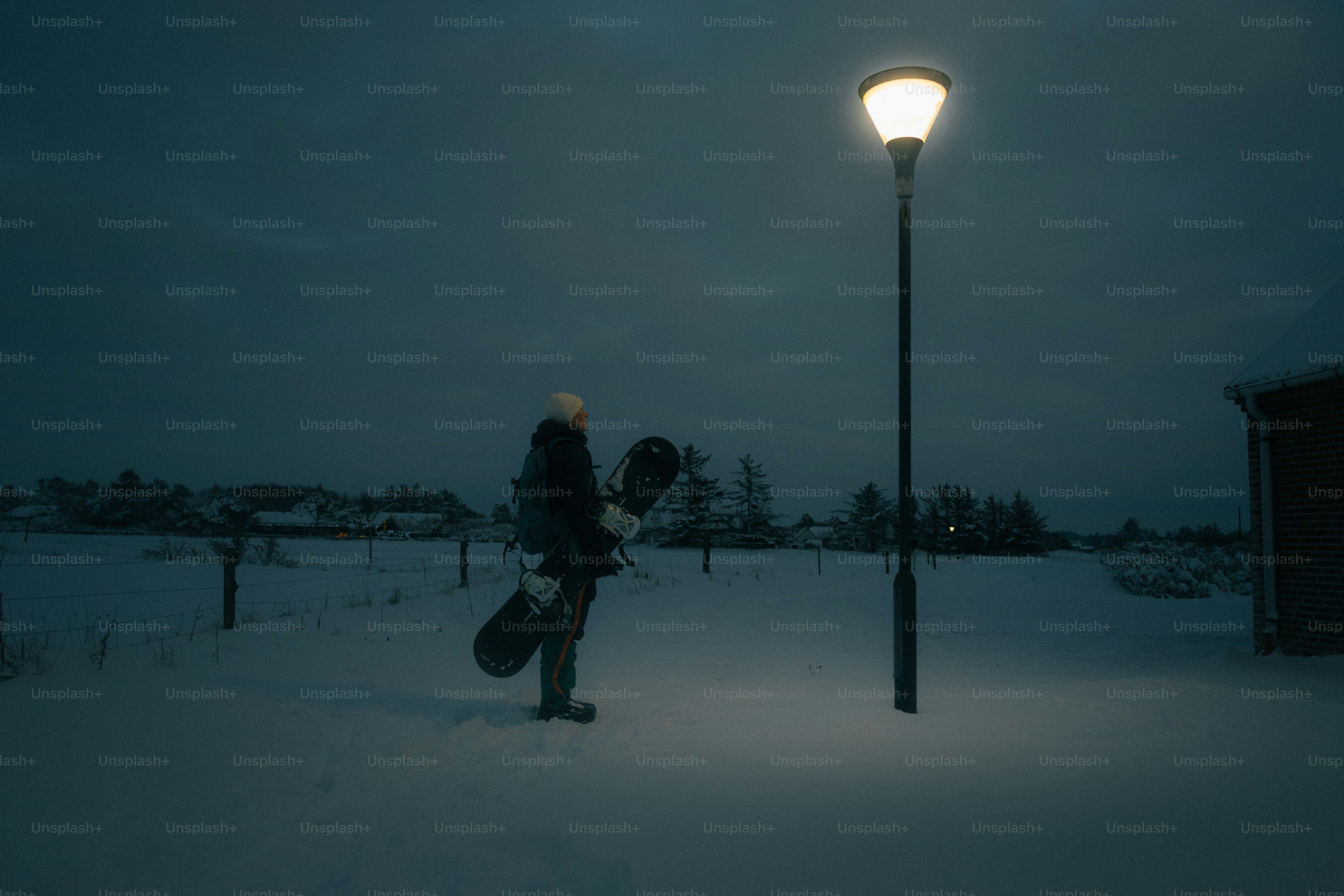 Person with snowboard stands under a streetlamp at night.