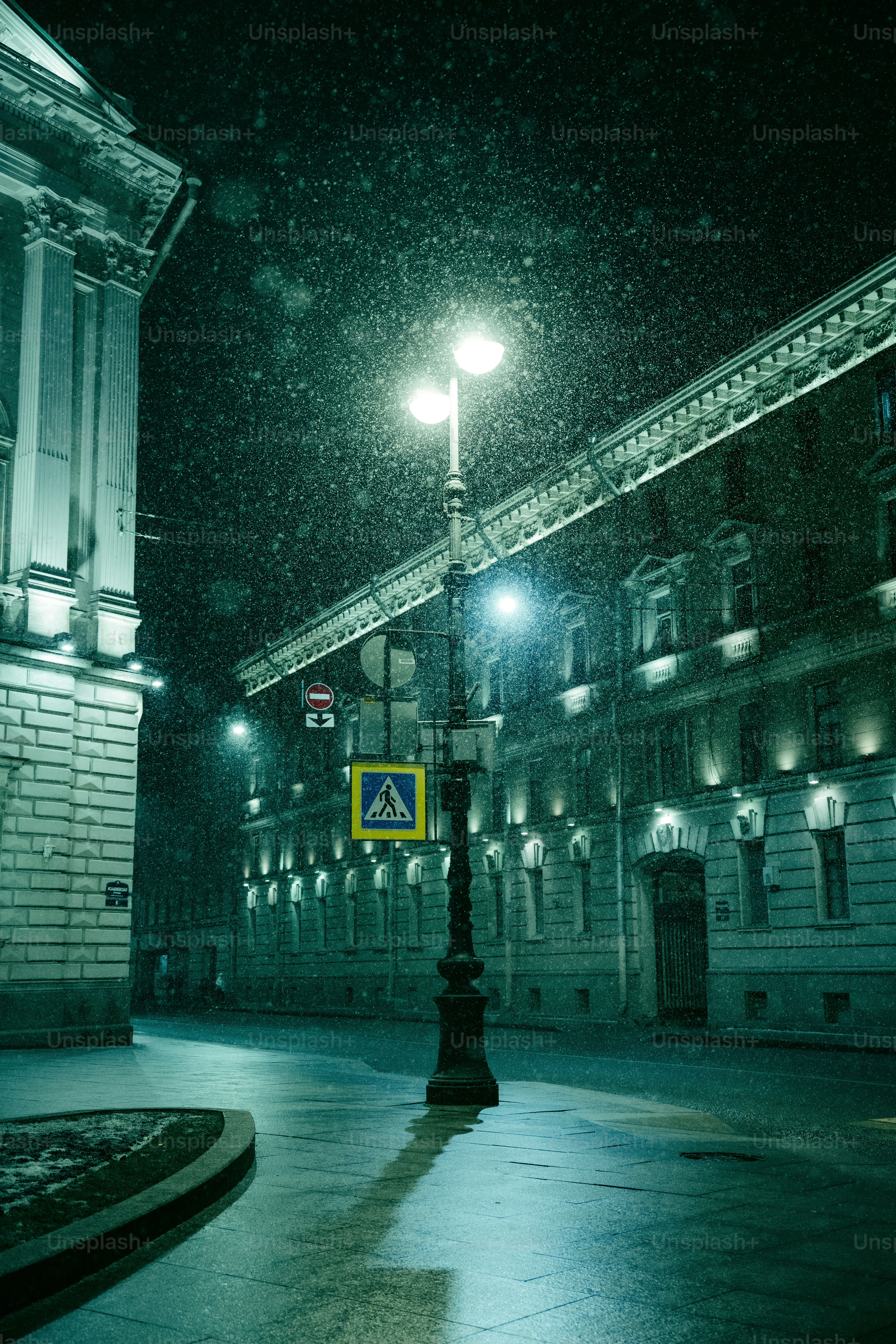 Street lamp illuminates wet street during snowfall at night.