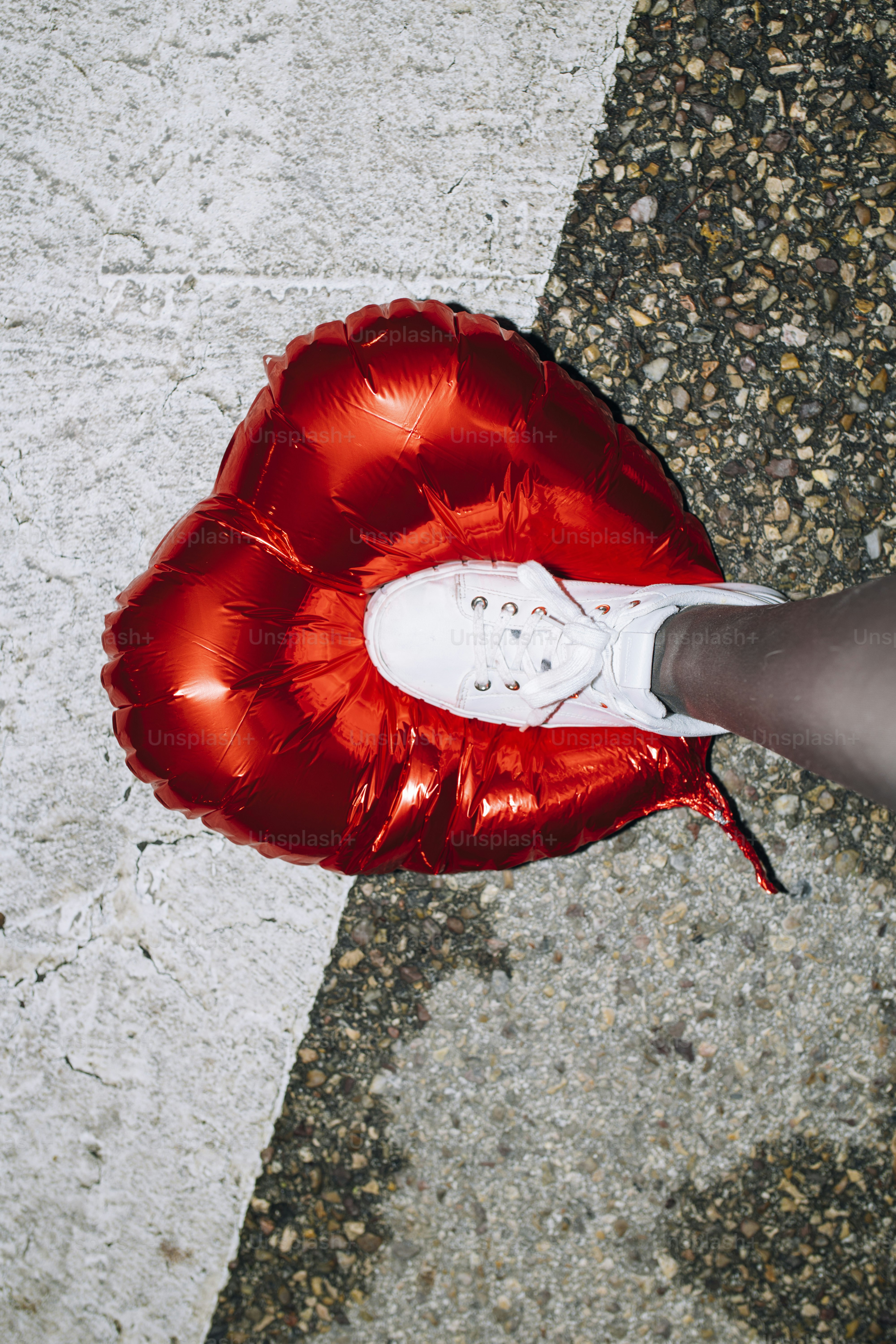 Ein weißer Turnschuh, der einen roten, herzförmigen Ballon zerquetscht.