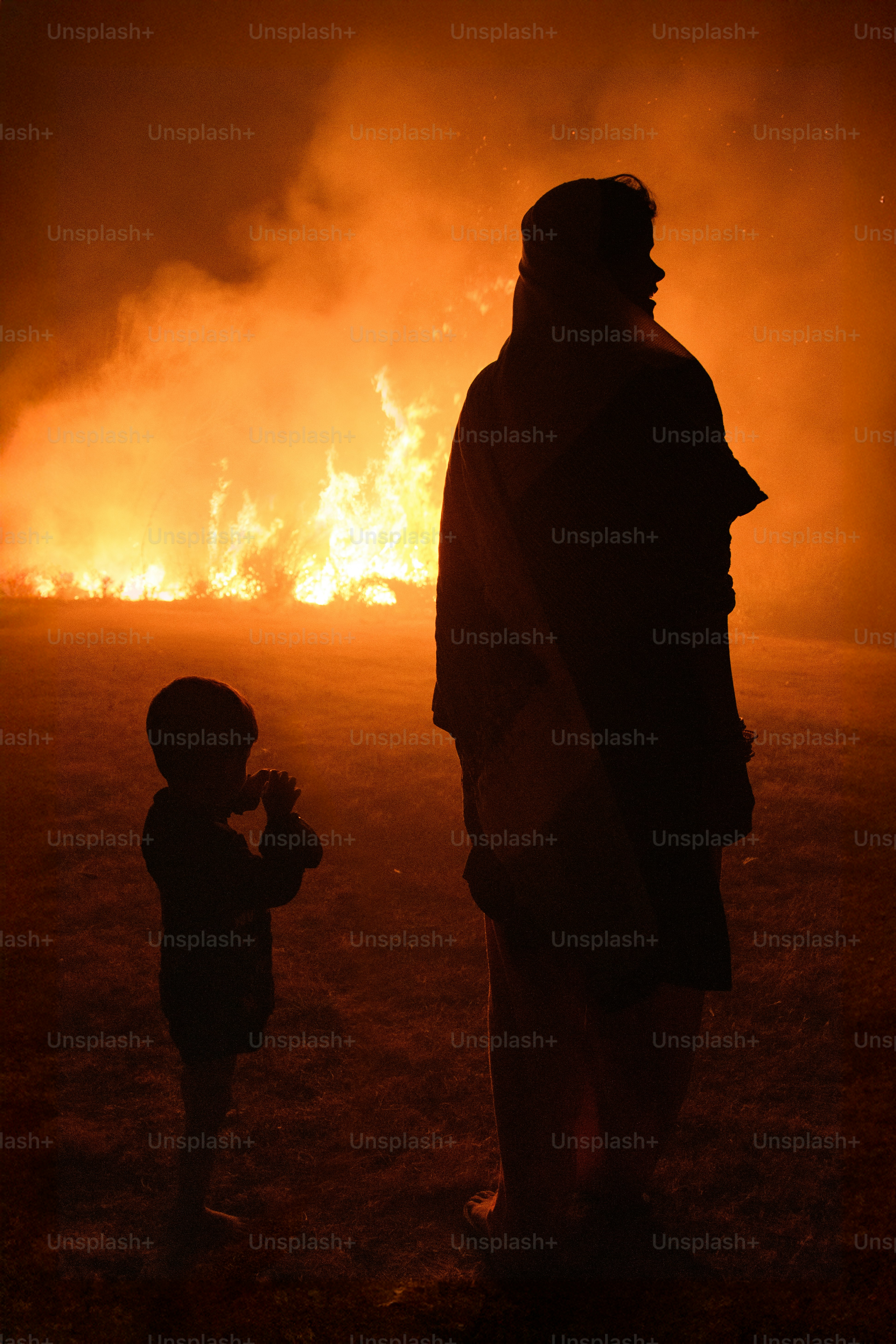 Two silhouetted figures watch a large bonfire at night.