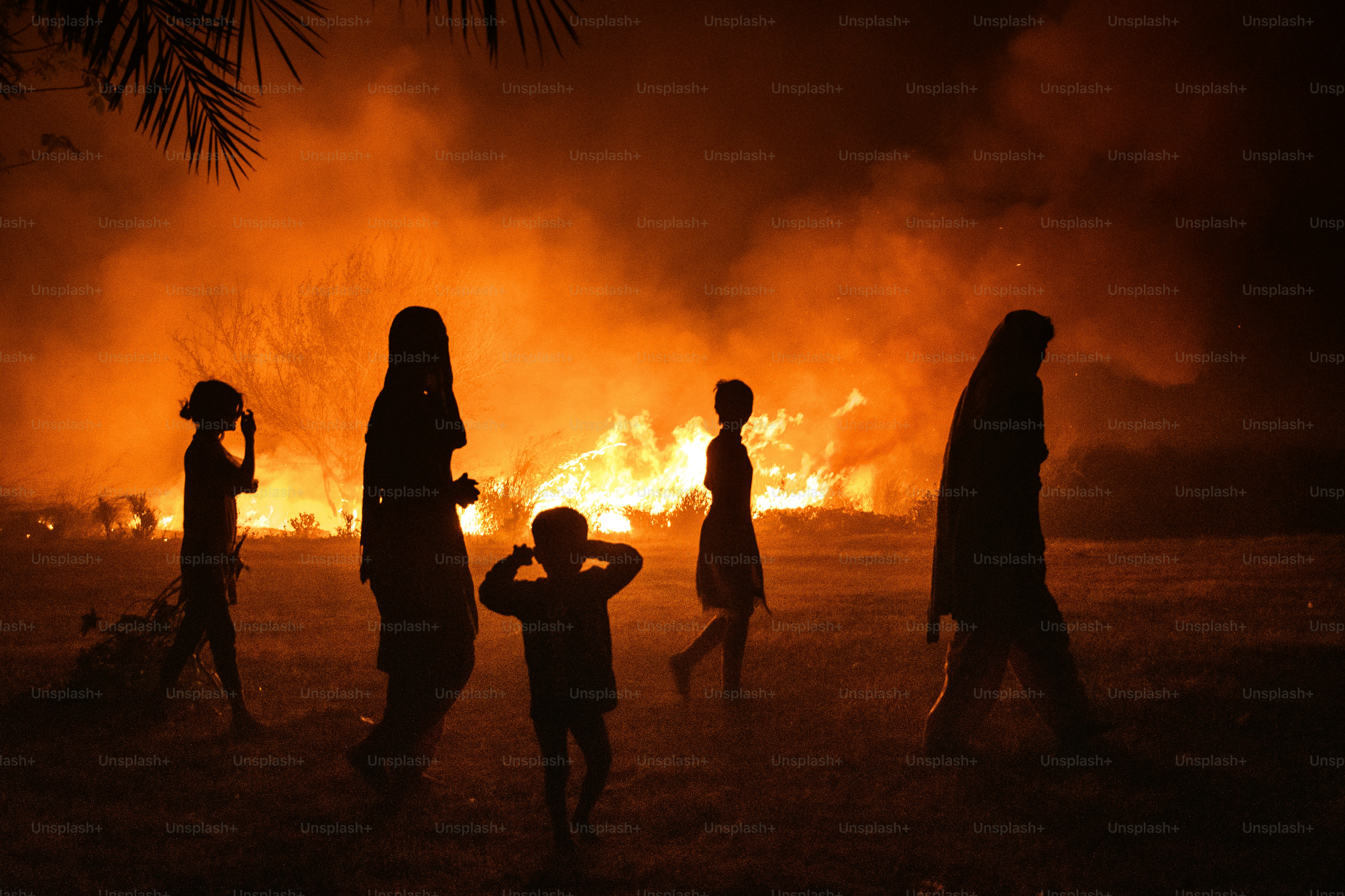 Silhouetted figures walk past a large fire at night.