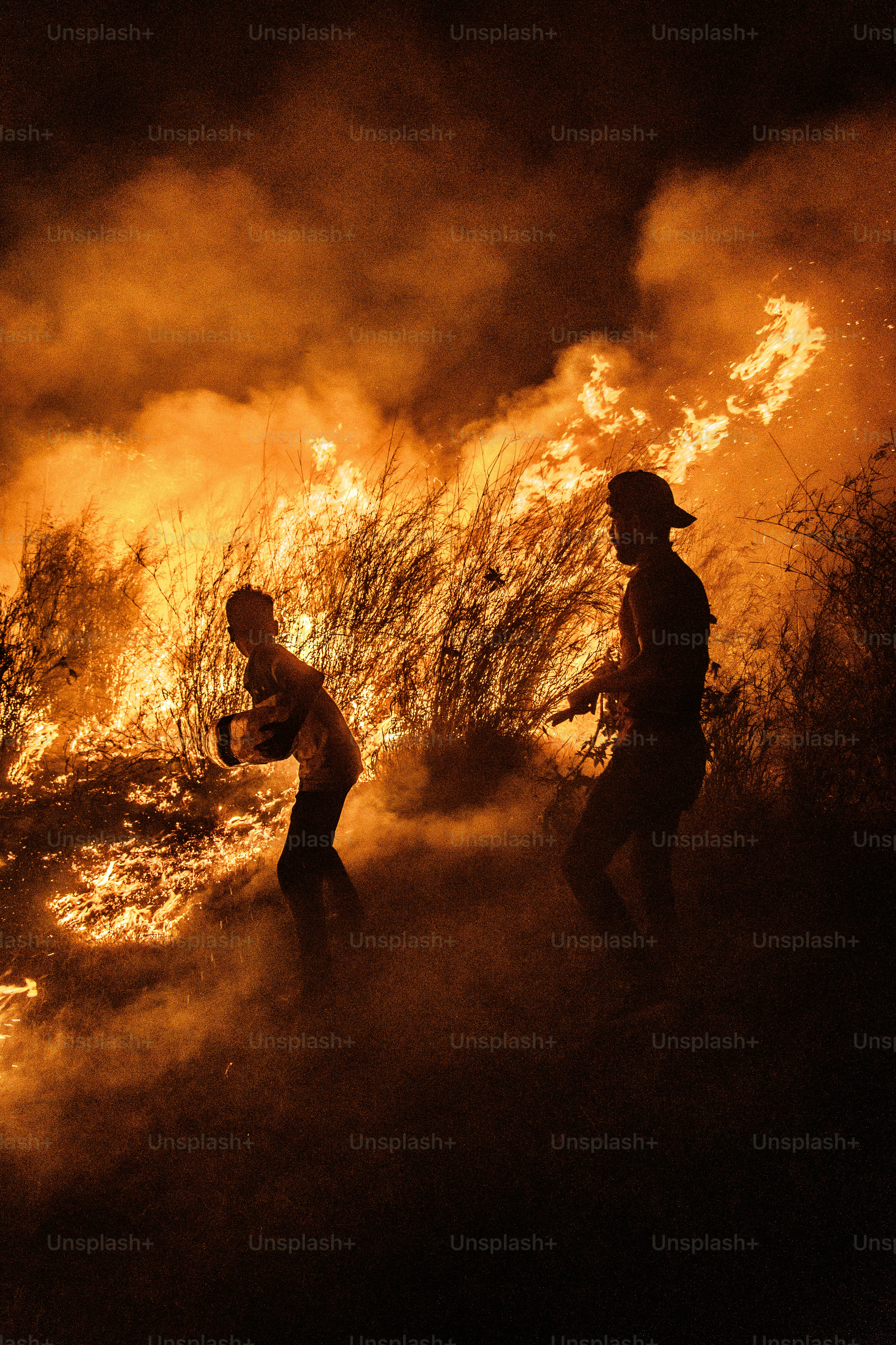 Two people walk through a large grass fire at night.