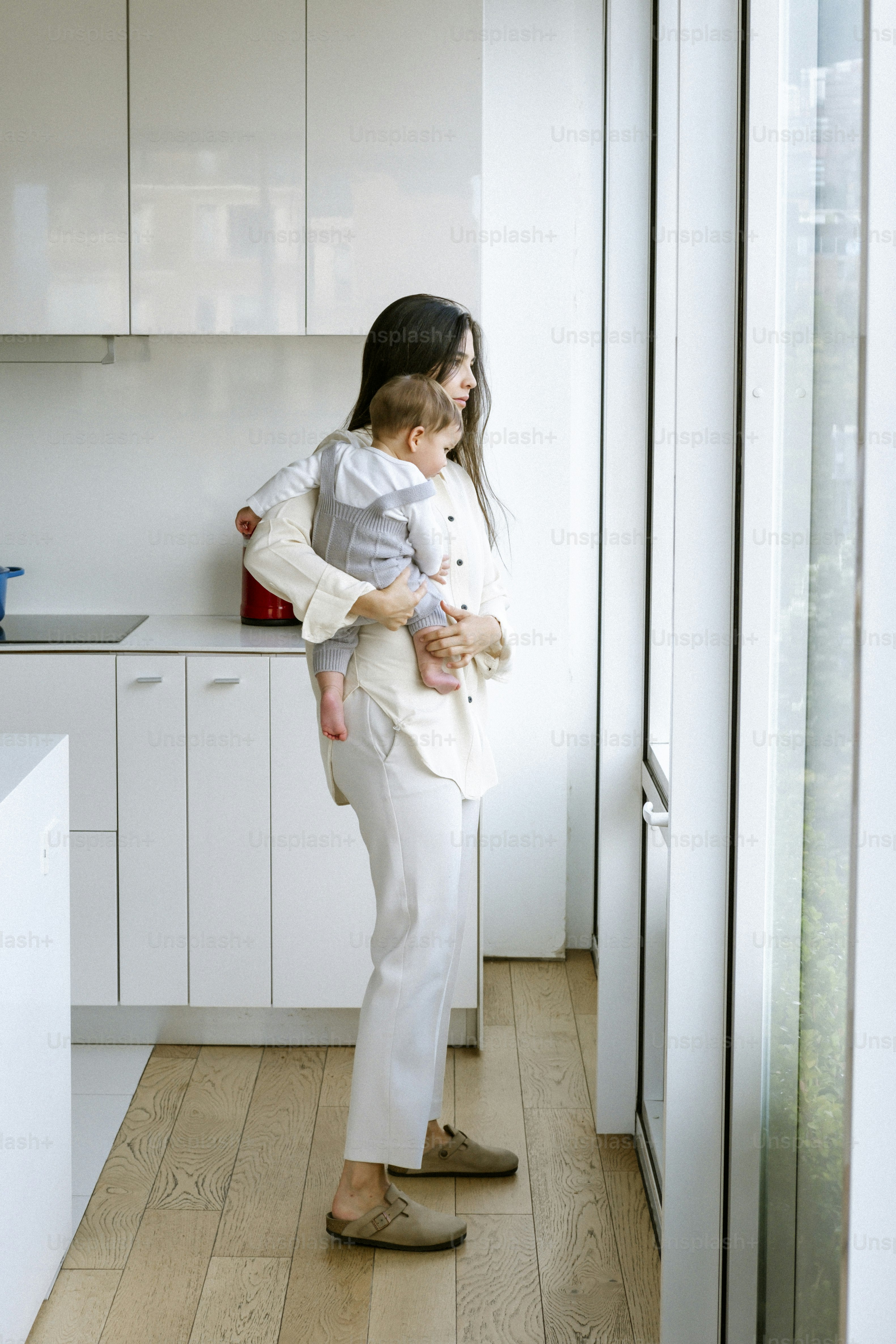 Mother holding baby looking out a large window.
