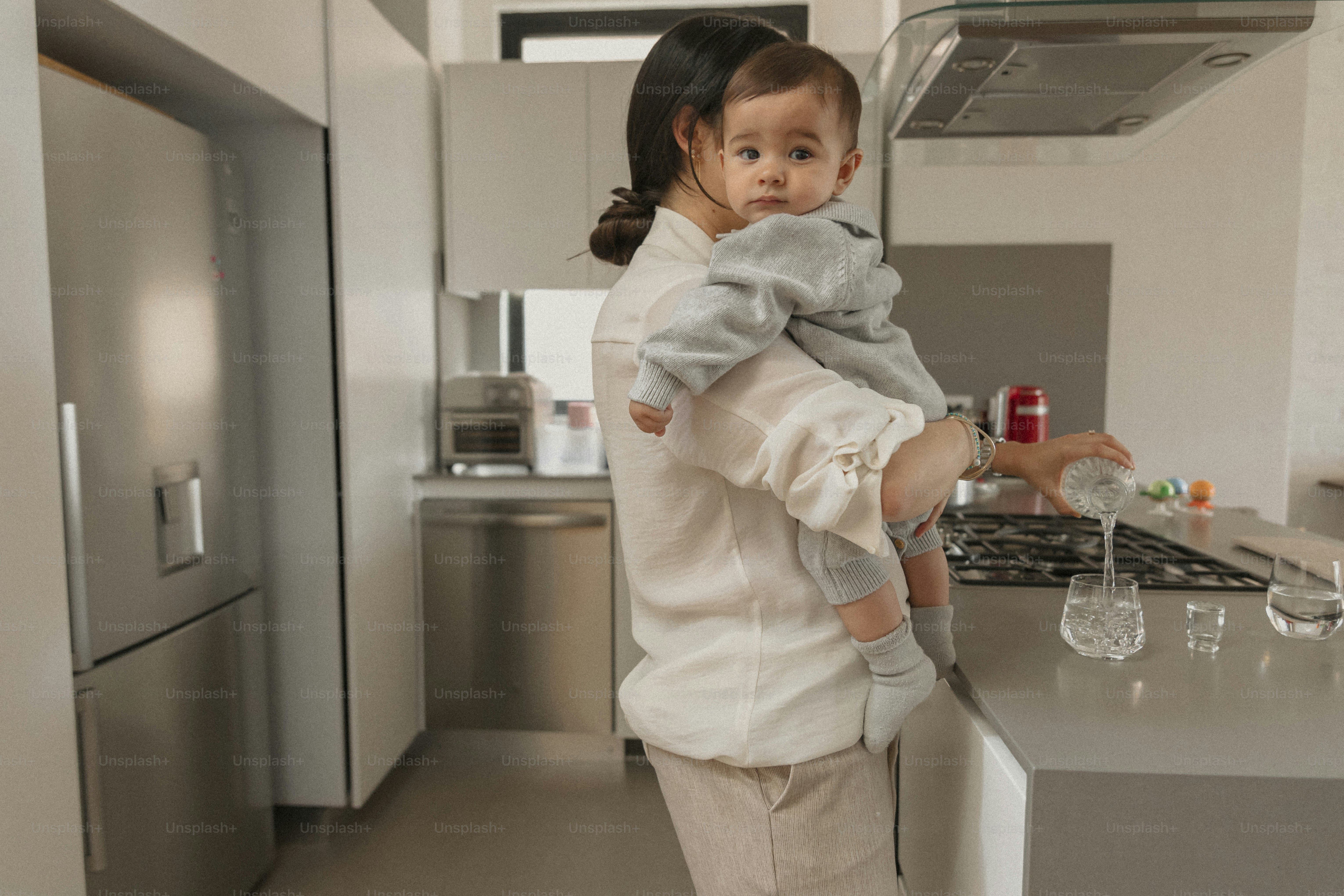 Mother holding baby while pouring water in kitchen.