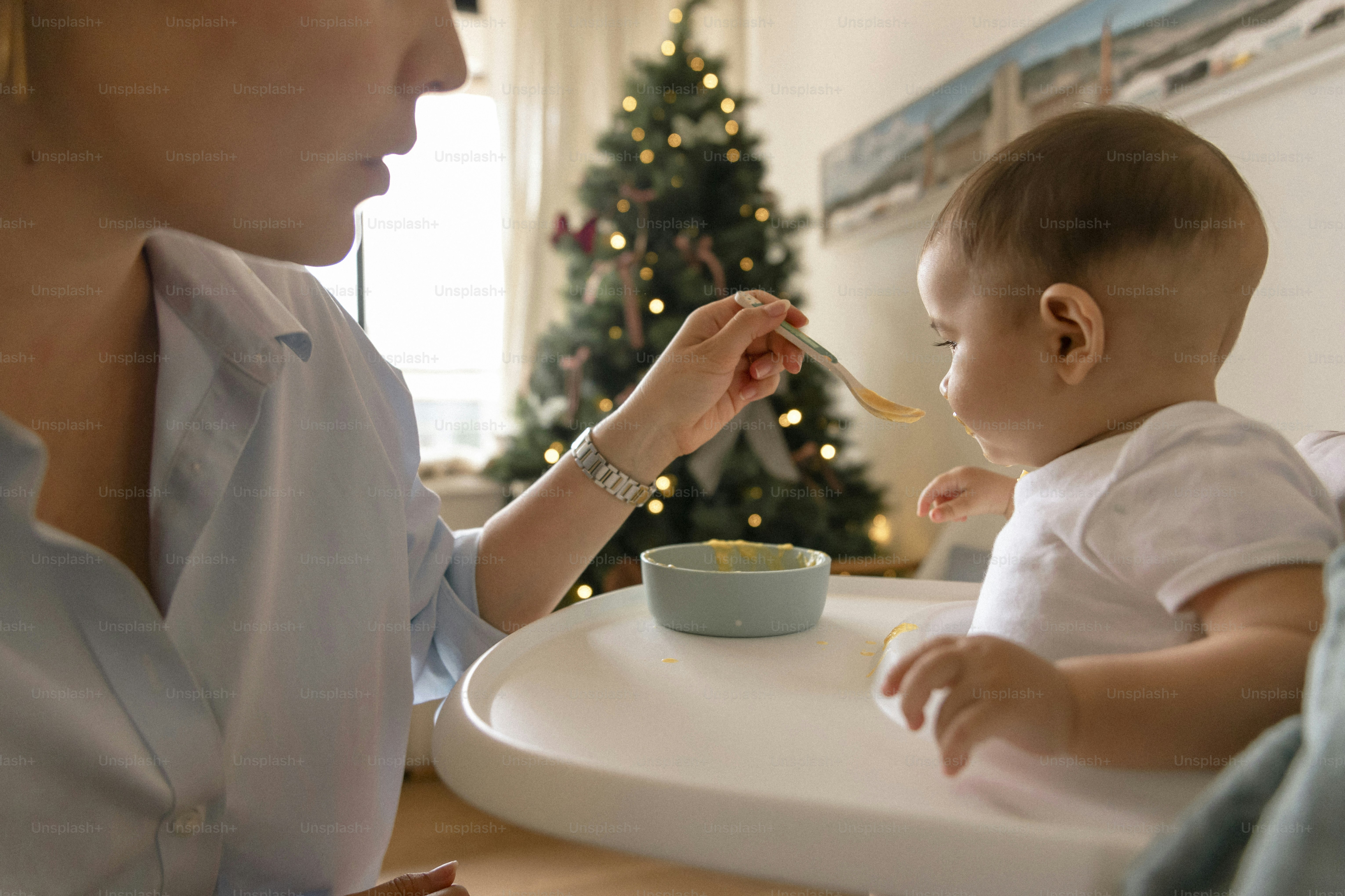 Mother feeding baby with a spoon near christmas tree.