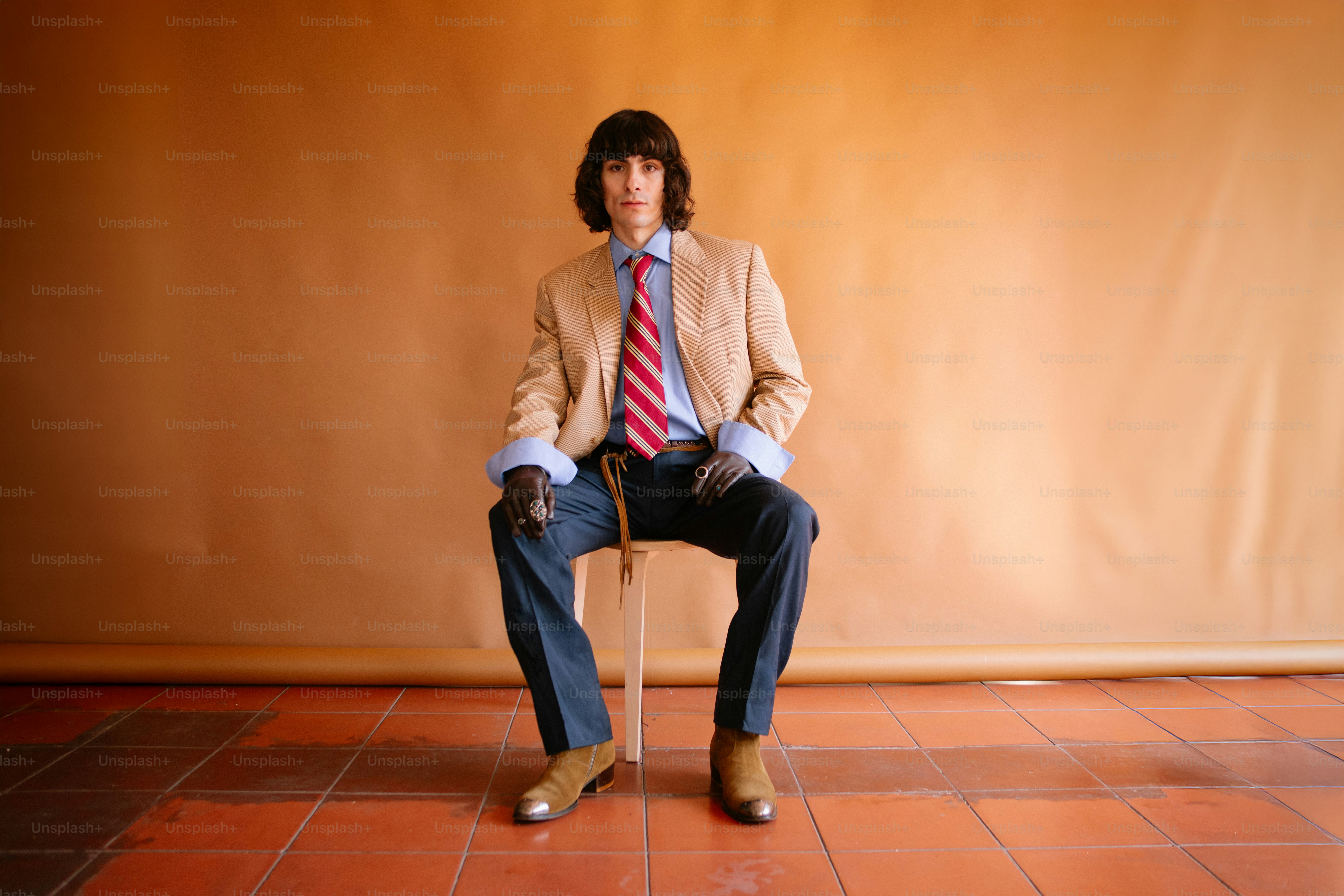 Uomo in abito elegante seduto su uno sgabello contro uno sfondo arancione.