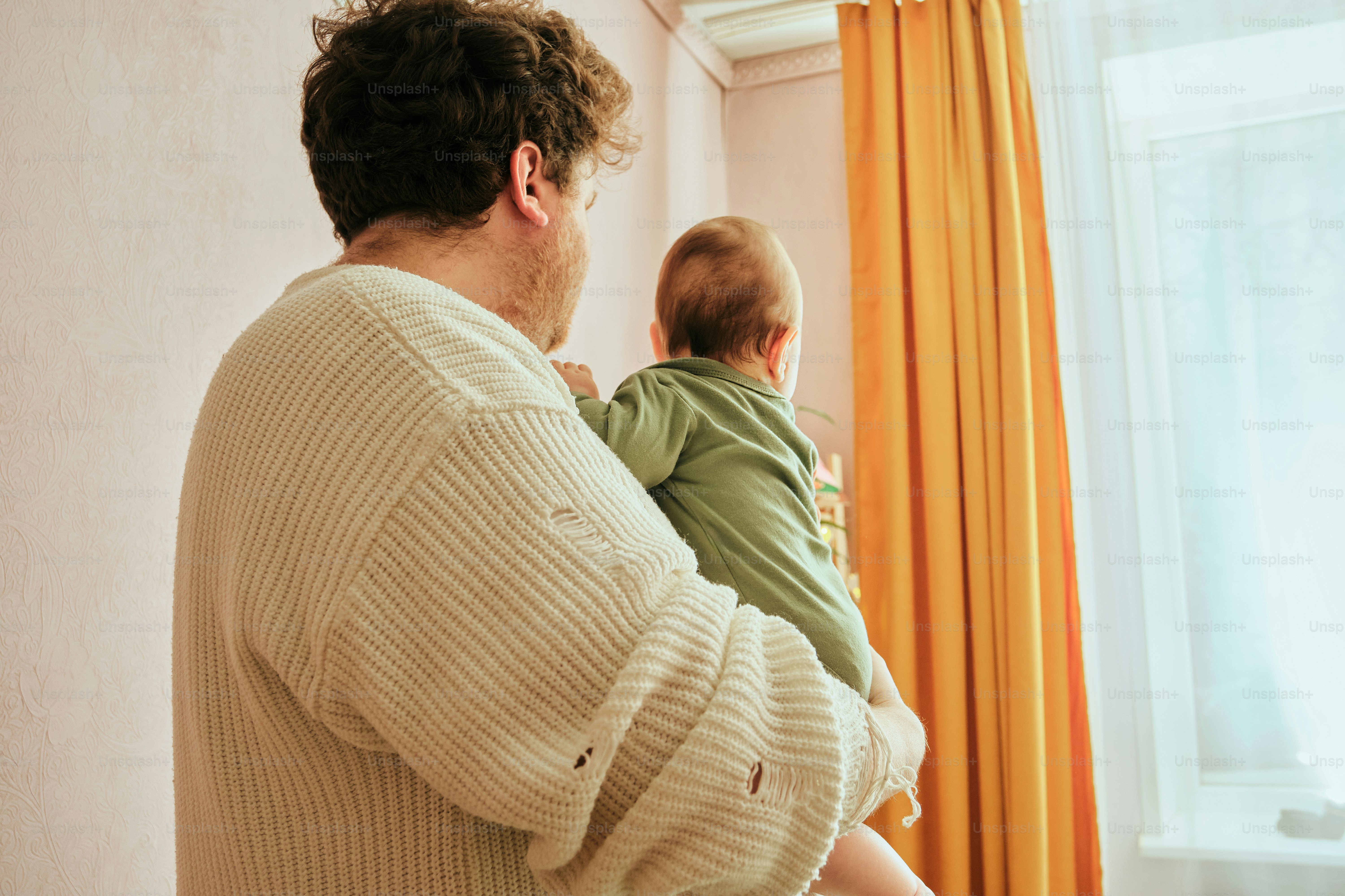 A man holds a baby near a window with curtains.