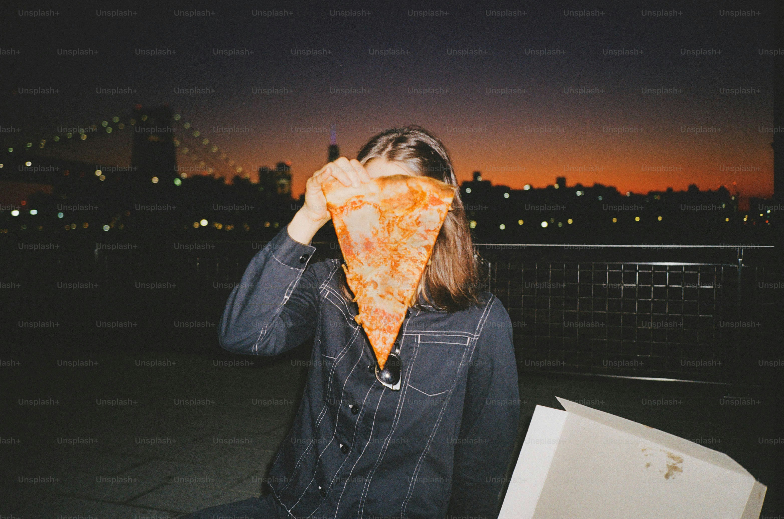 Person holding pizza slice in front of face at dusk