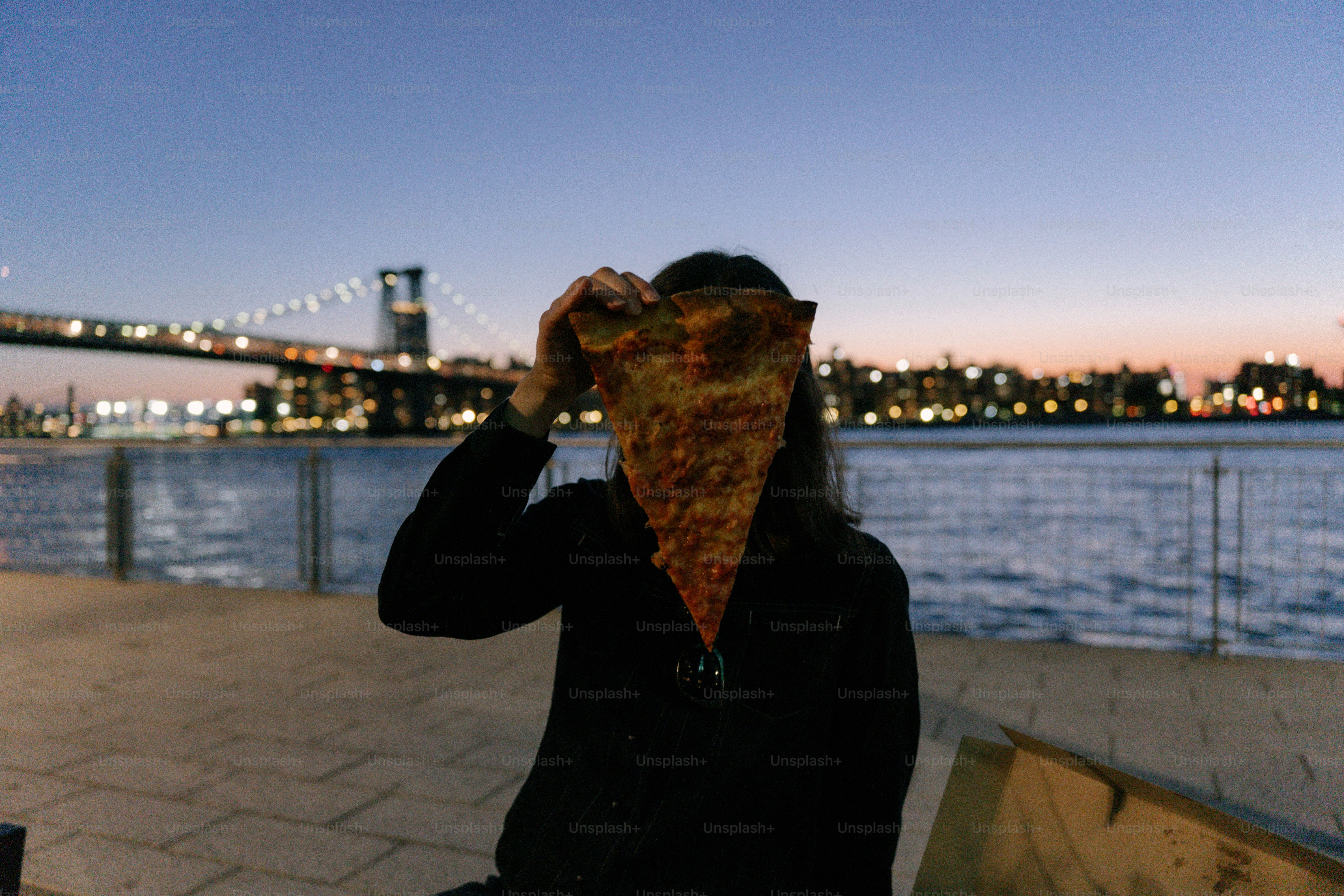 사람이 피자 조각을 얼굴 위로 들고 있으며, 배경은 도시의 불빛 아래서
