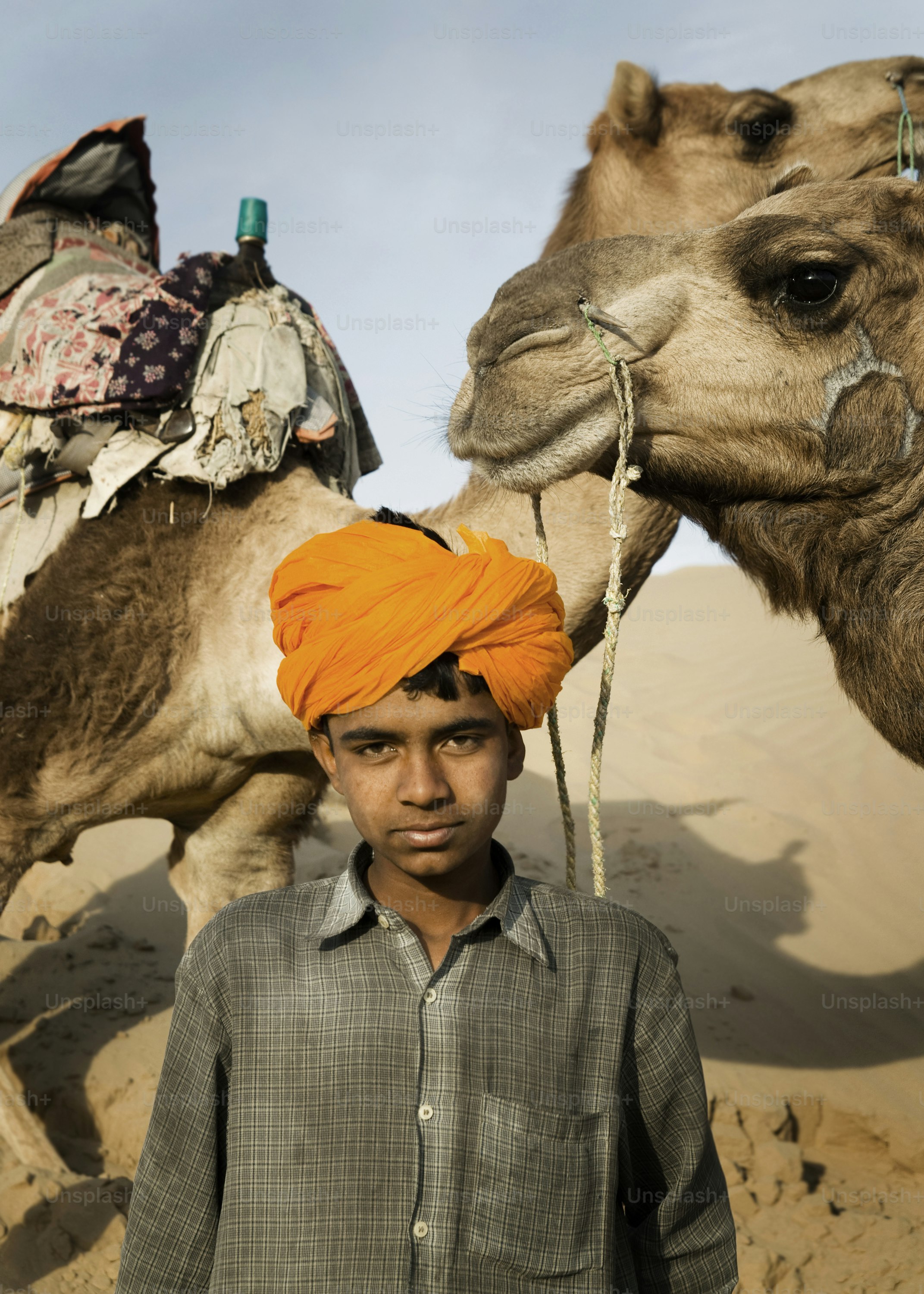 Young boy with orange turban and camel in desert.