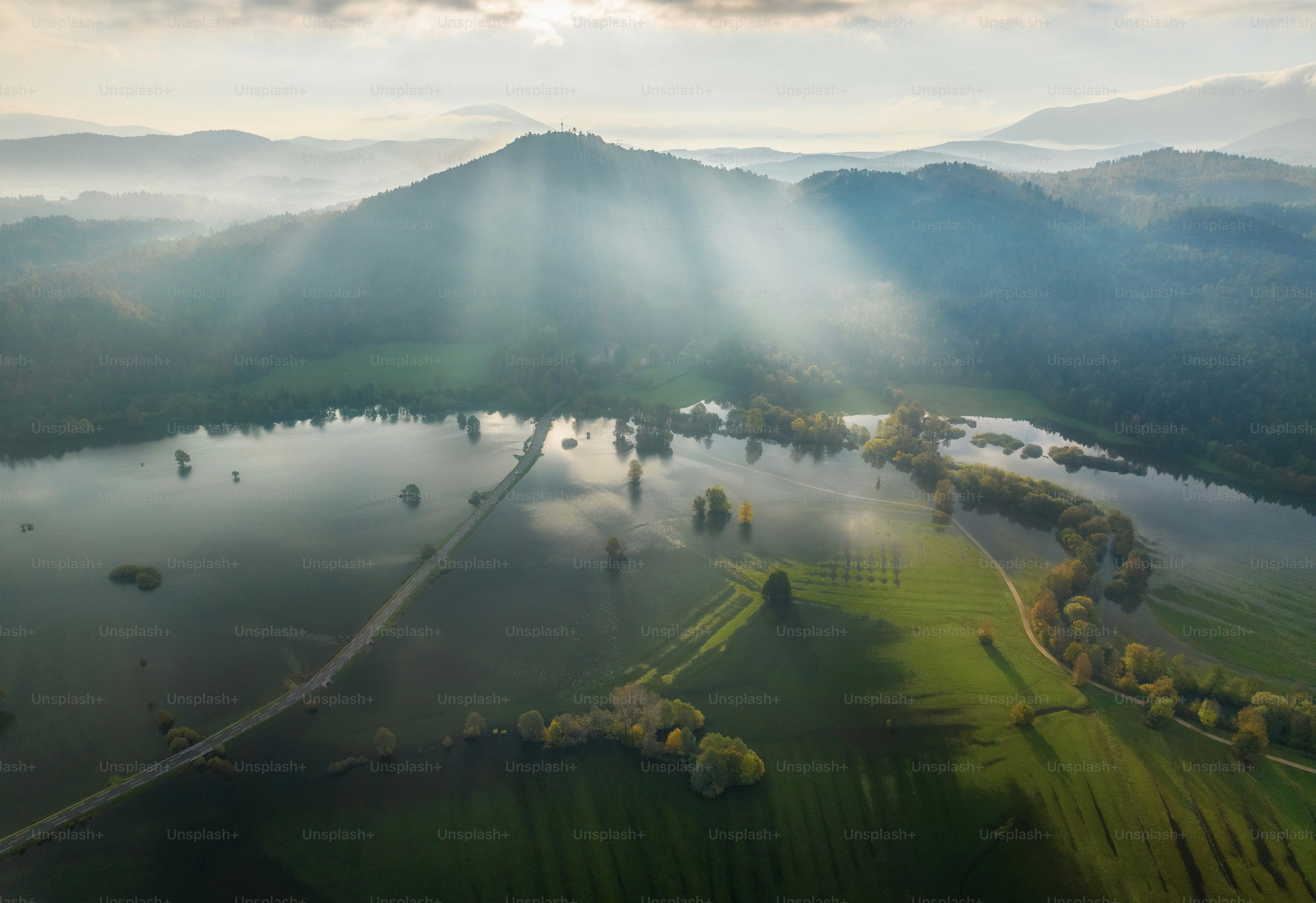 Sonnenstrahlen scheinen durch Nebel über überfluteten Feldern und Hügeln.